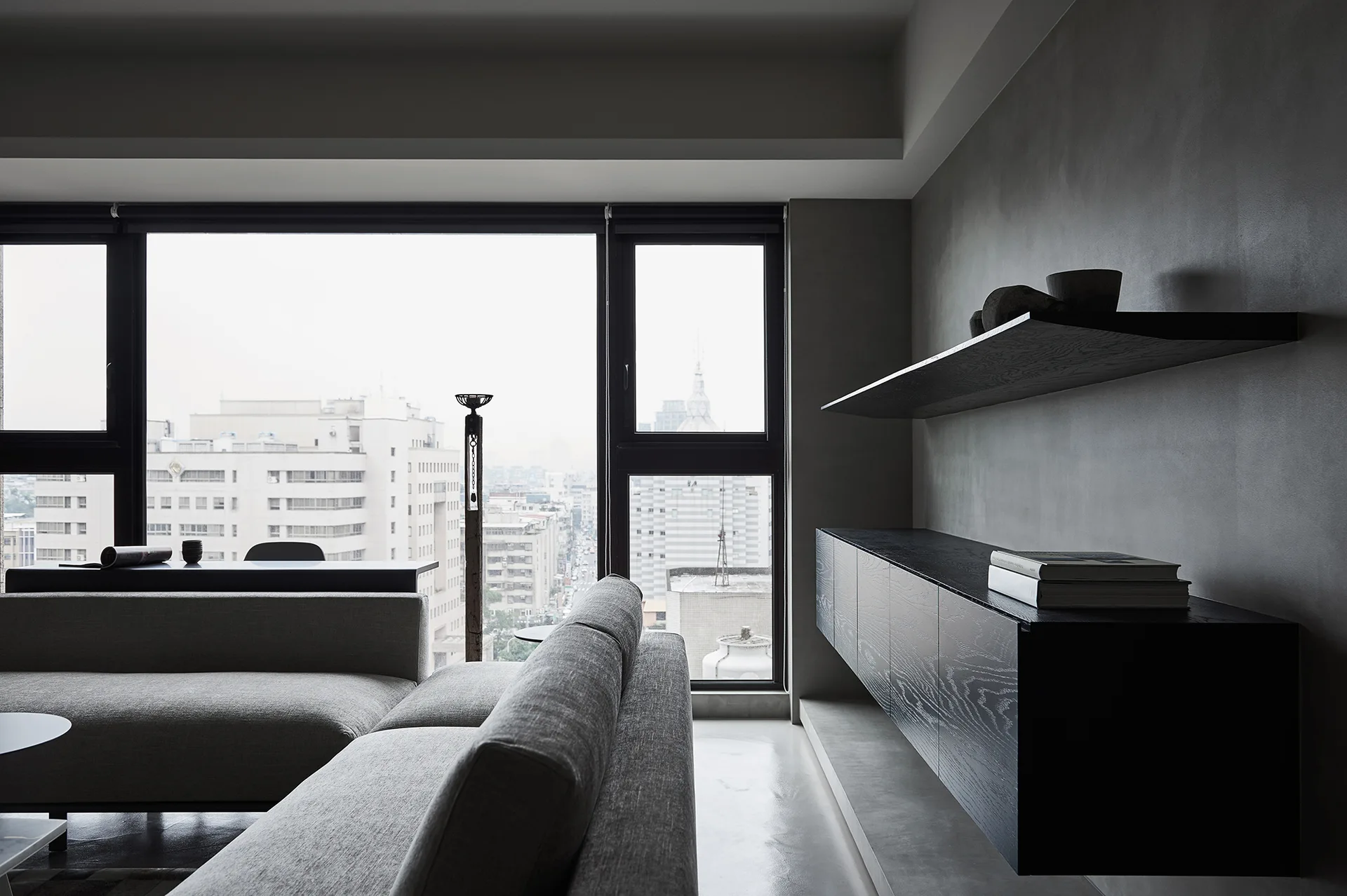 A minimalist living room featuring a grey sectional sofa, a black wood floating cabinet on a grey wall, and a large window overlooking the city.