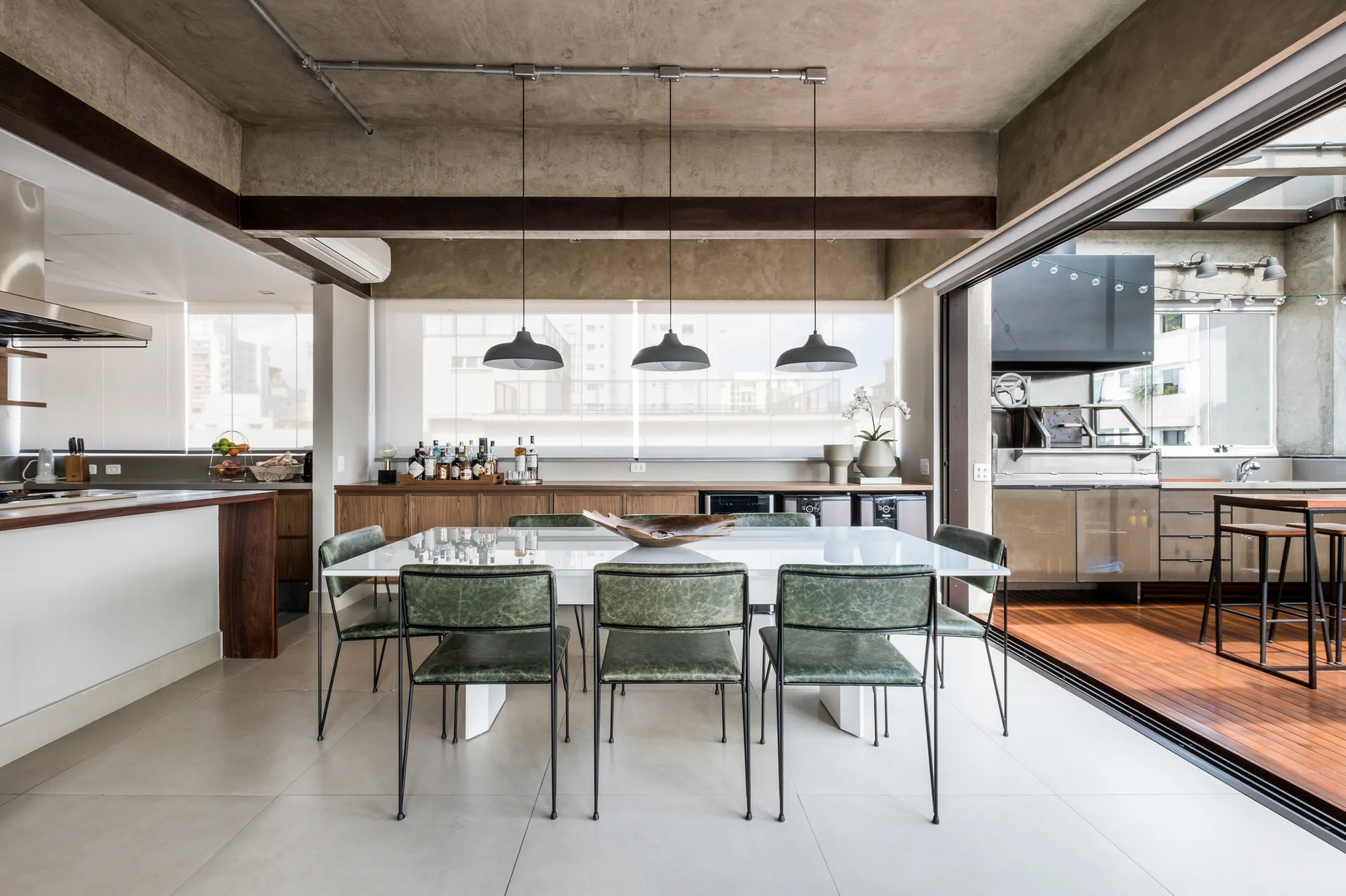 A modern dining room features a white table with green chairs under an exposed concrete ceiling, next to a sliding glass door opening to a wooden deck.