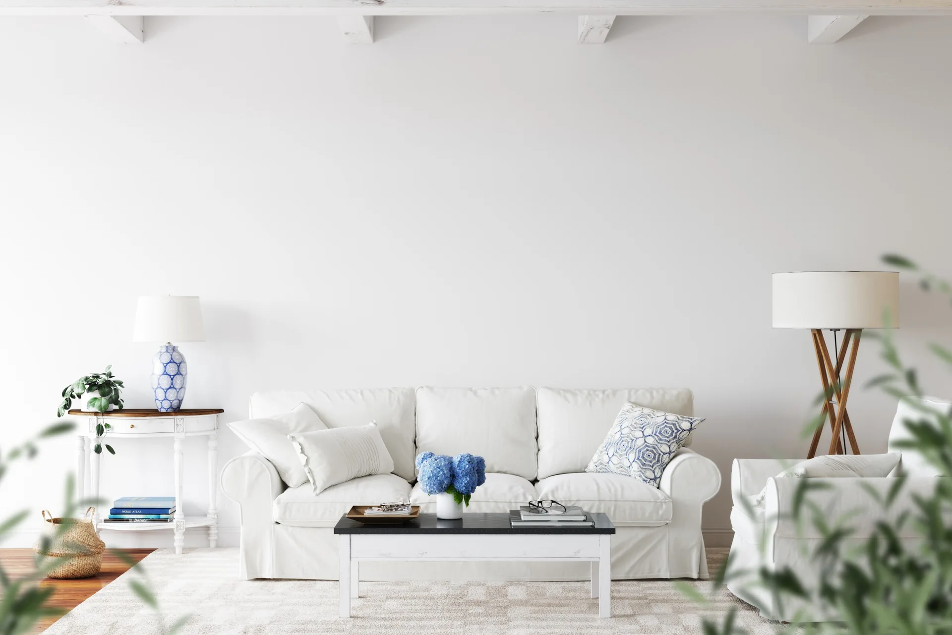 A bright white living room featuring a slipcovered sofa, a blue-and-white patterned lamp on a console table, and exposed ceiling beams.