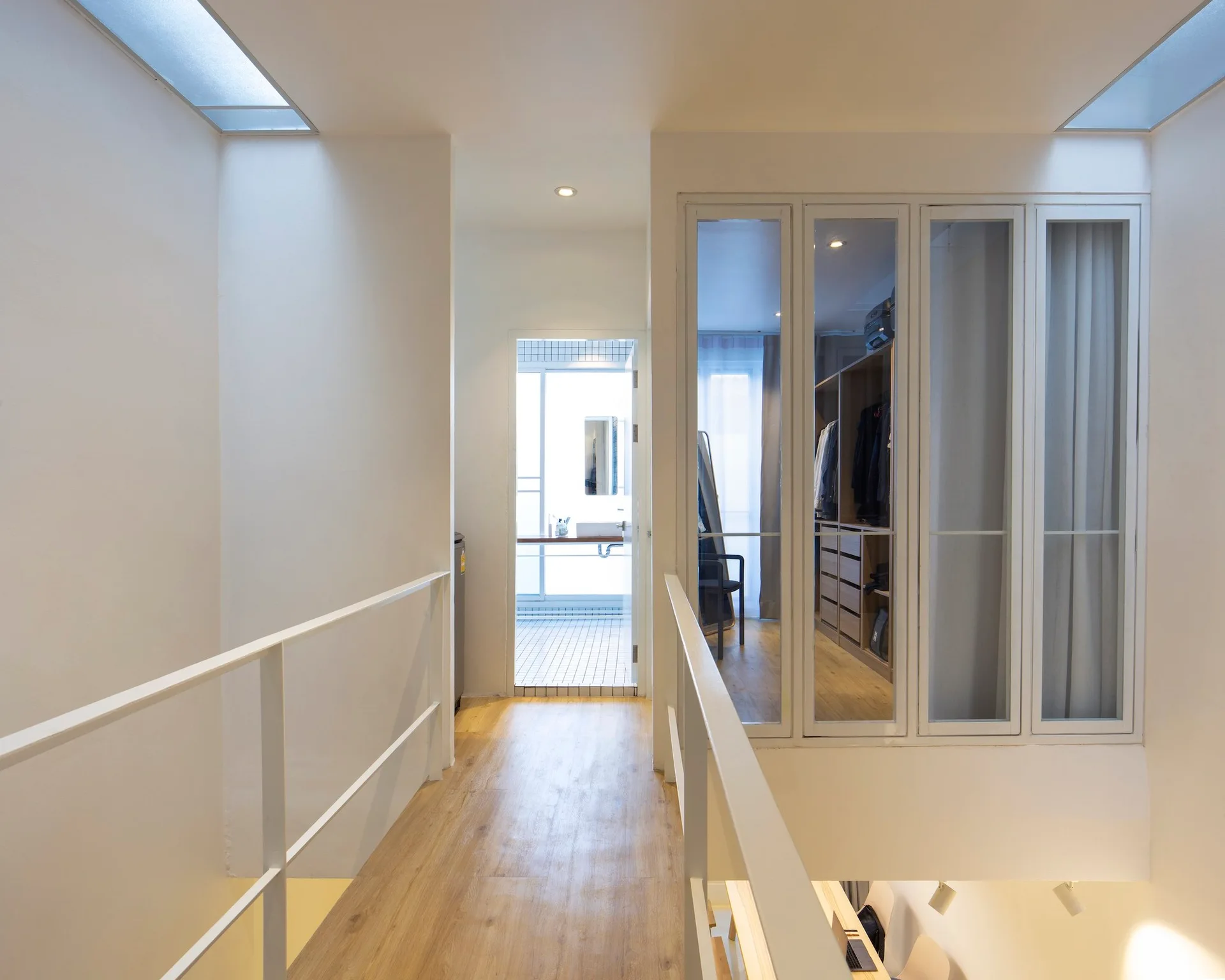 A bright modern bridge hallway with white metal railings, light wood flooring, skylights, and a glass-enclosed walk-in closet.