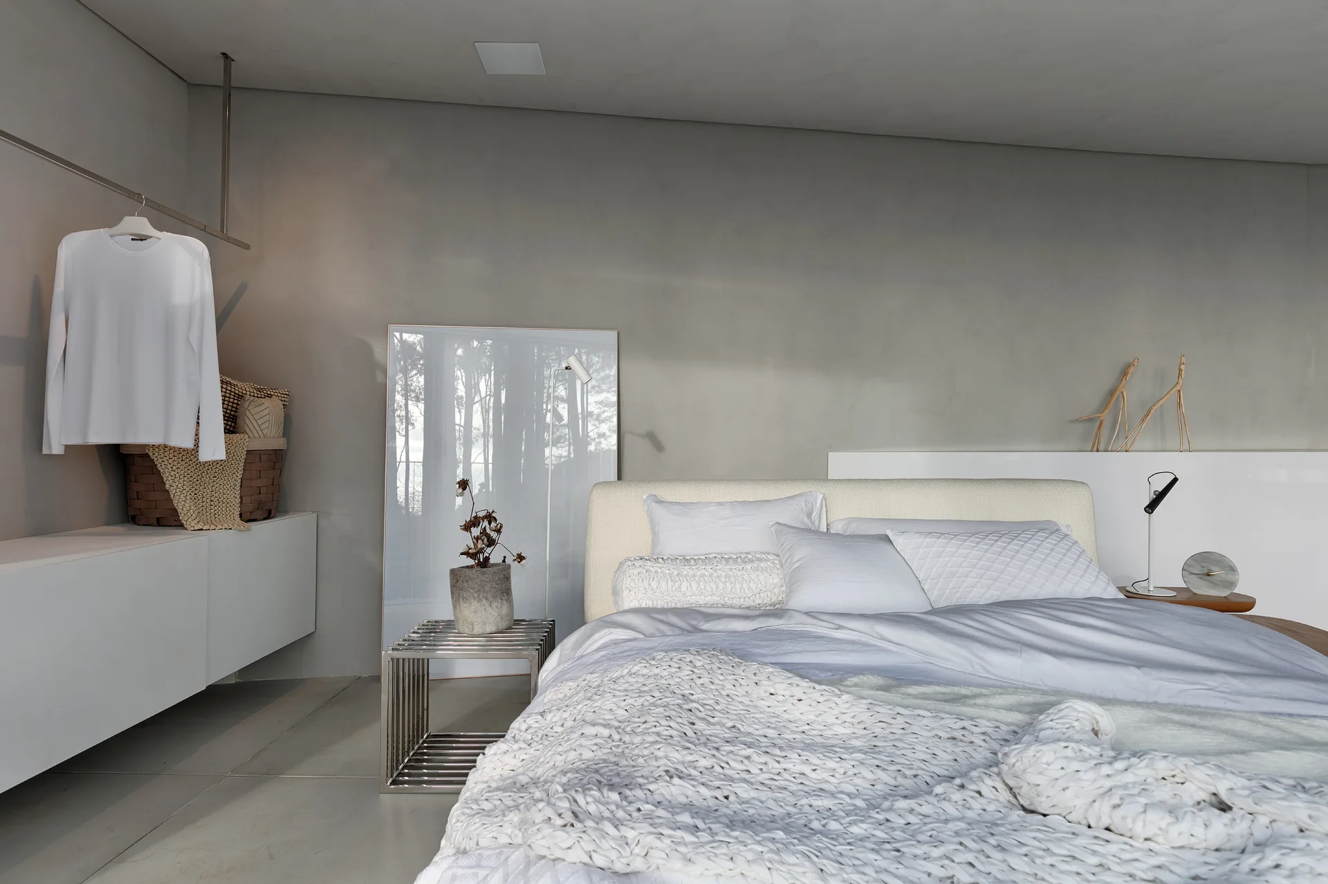 A minimalist bedroom featuring a bed with white bedding and a chunky knit throw, a metal grid side table, and smooth grey plaster walls.
