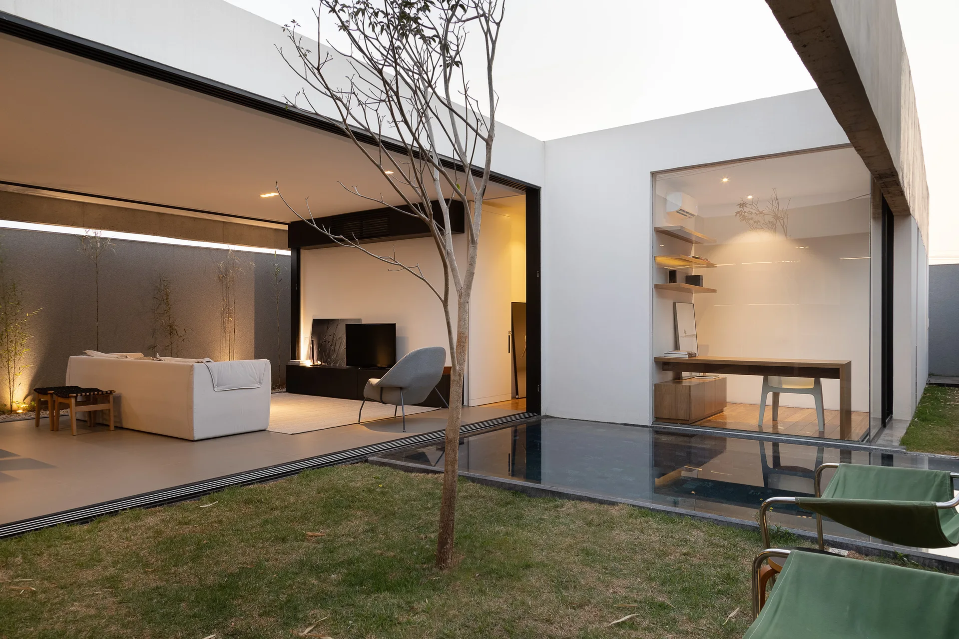 A minimalist home features an open-sided living room and a glass-enclosed office overlooking a grassy courtyard with a small pool.