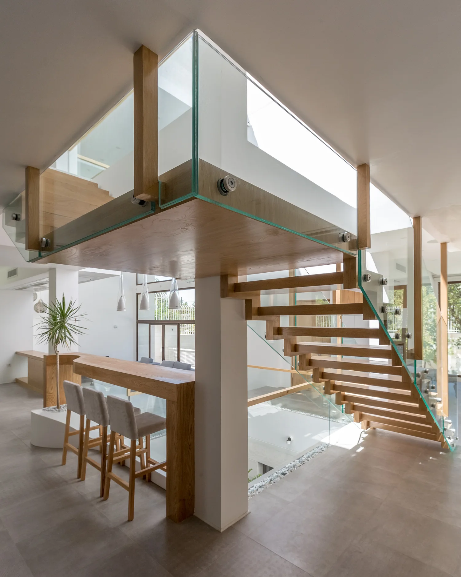 A modern interior features a floating wooden staircase with glass railings built over a wooden bar counter with three upholstered stools.