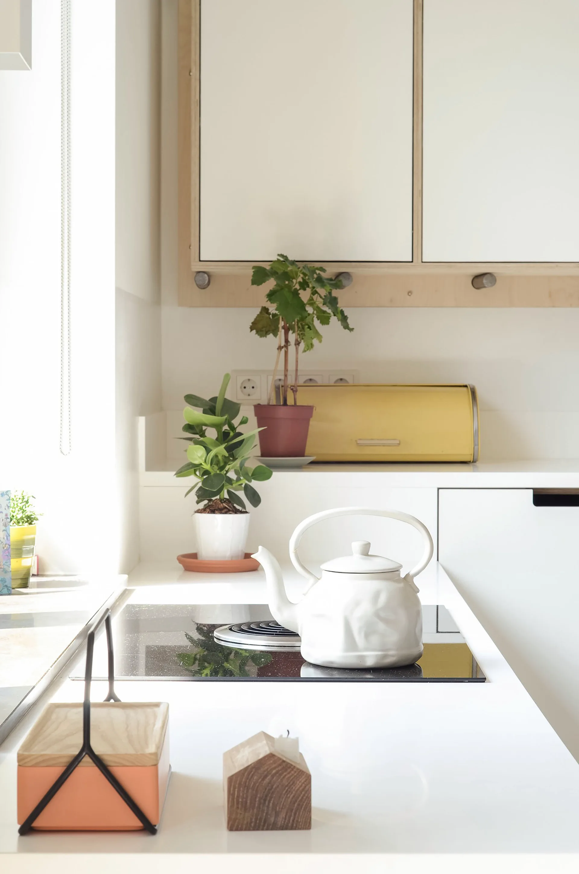 A bright kitchen counter features a black glass stovetop with a white kettle, wood-trimmed white cabinets, two small potted plants, and a yellow breadbox.