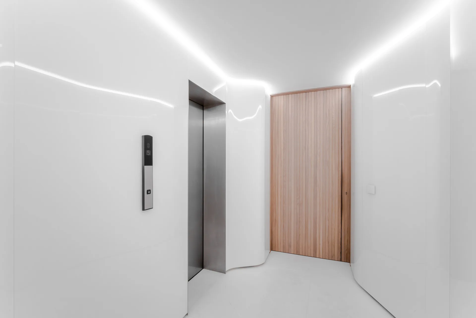 A minimalist hallway features high-gloss curved white walls, a stainless steel elevator, integrated cove lighting, and a vertical slatted wood door.