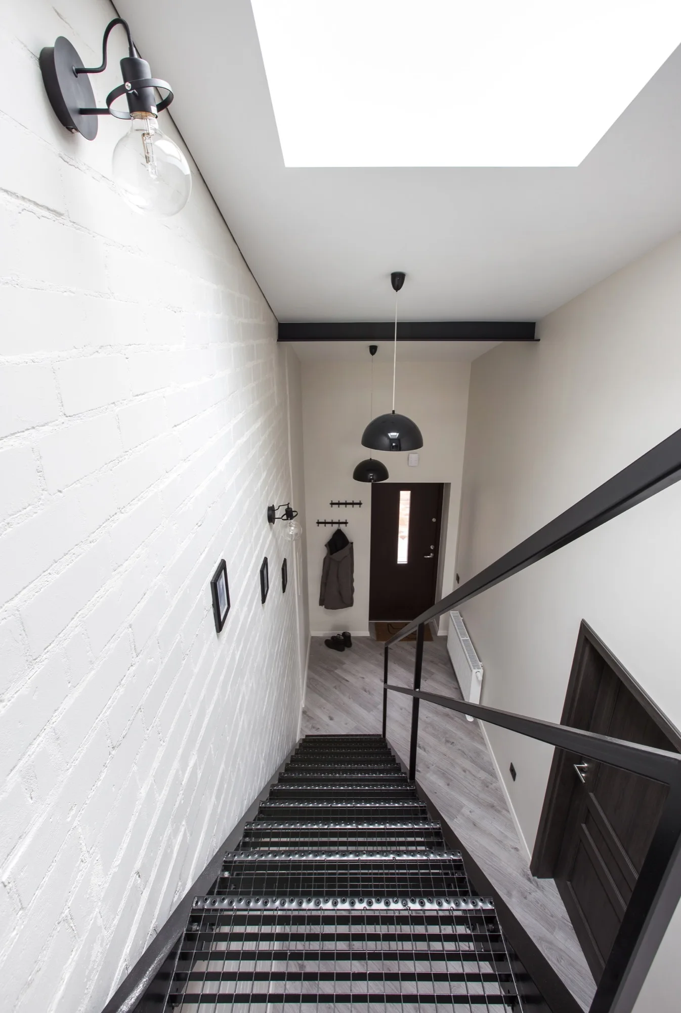 A view looking down a black metal industrial staircase with open treads next to a white painted brick wall leading to a grey herringbone floor entryway.