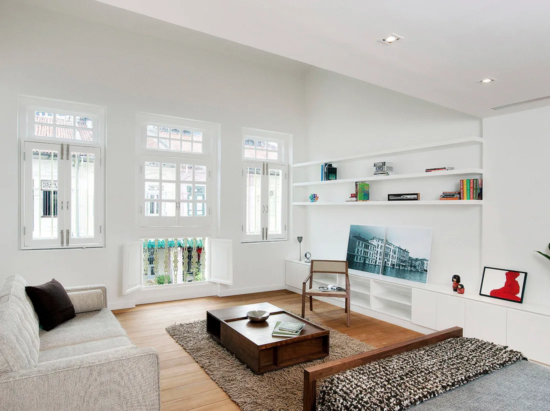 A minimalist living room featuring a gray fabric sofa, a low wooden coffee table, white shuttered windows, and integrated wall shelving.