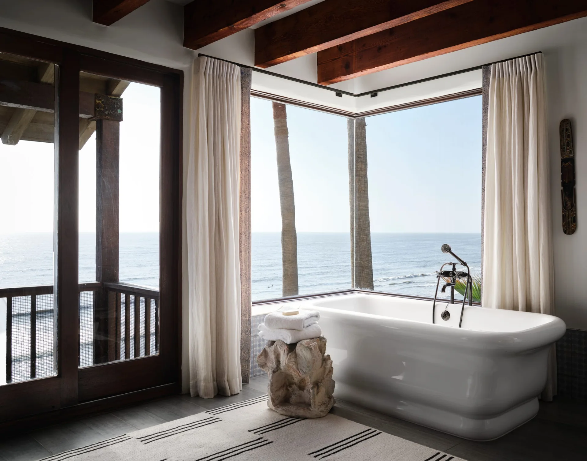 A bright bathroom features a white freestanding bathtub next to a large corner window showing an ocean view, with dark wood ceiling beams above.