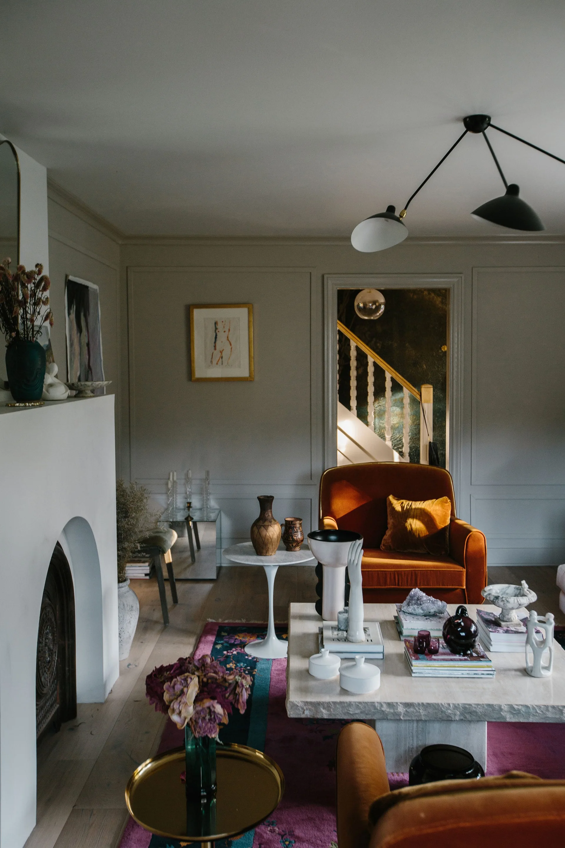 An eclectic living room features a bright orange velvet armchair, a low stone coffee table, and a minimalist white arched fireplace against gray paneled walls.
