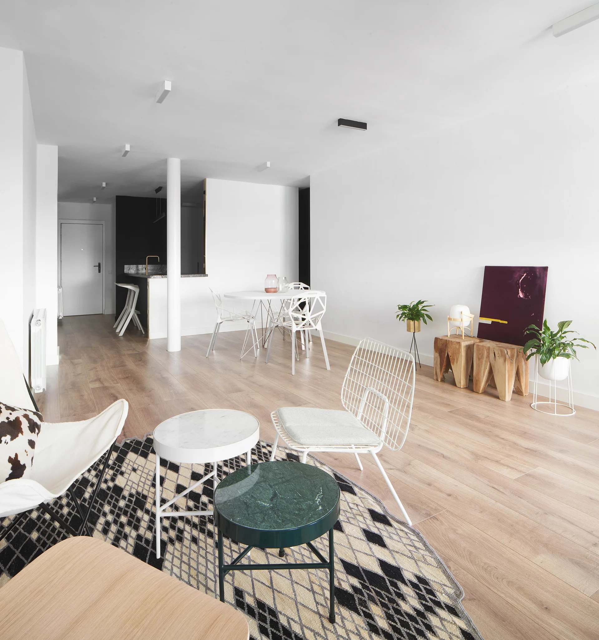 A minimalist open-concept living area with light wood floors, a bold geometric rug, a white wireframe chair, and a round dining table in the background.