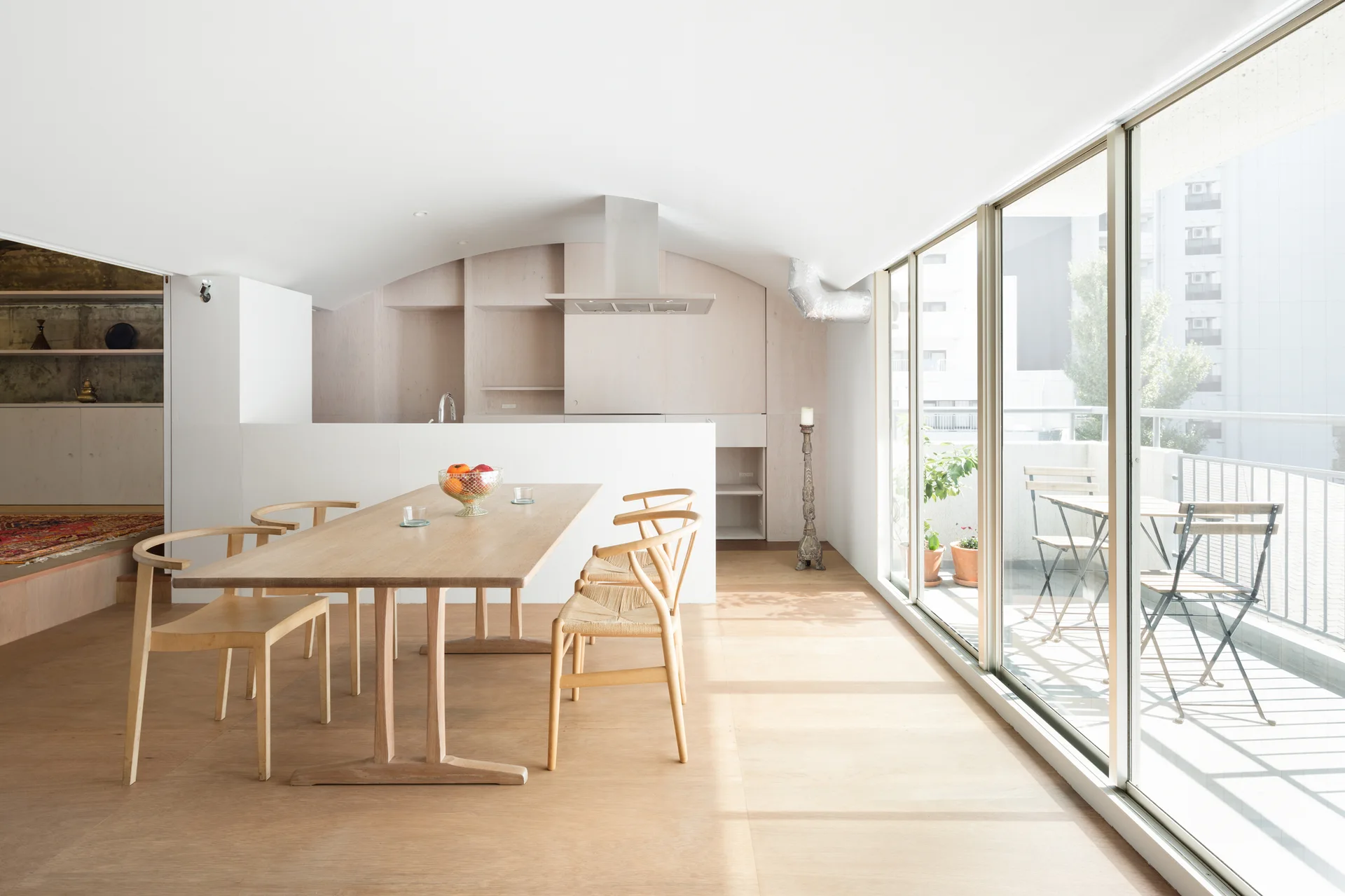 A minimalist dining space features a light wood table and woven chairs next to floor-to-ceiling glass doors opening onto a balcony.