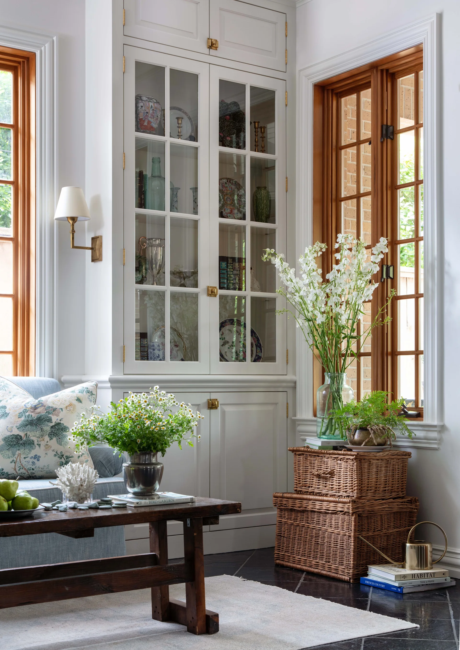 A white built-in glass-door cabinet stands between two warm wood-trimmed windows, with stacked wicker baskets and a low wooden coffee table nearby.