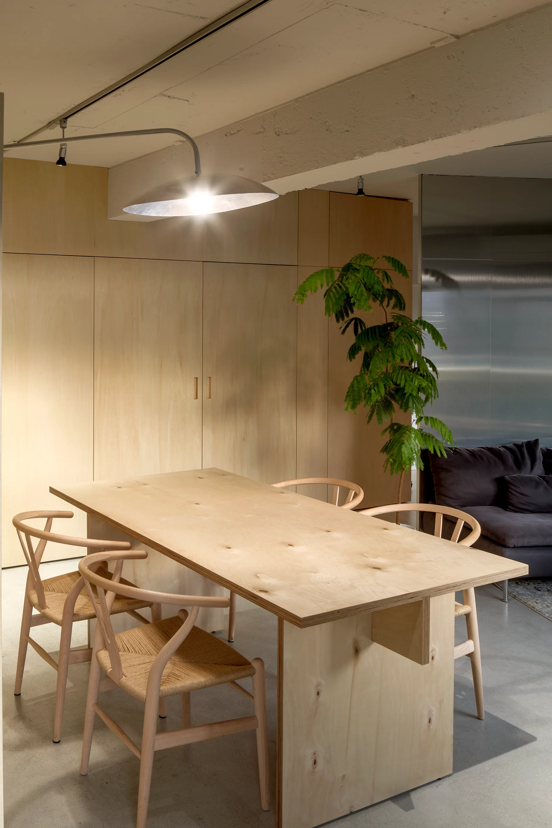 A minimalist dining room features a custom plywood table, woven-seat wood chairs, seamless wooden cabinetry, and a large metal dome pendant light.