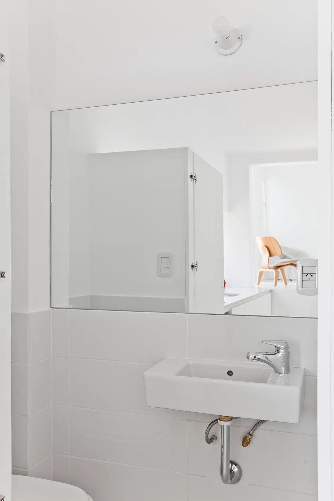 A minimalist white powder room featuring a wall-mounted ceramic sink, a large frameless mirror, and clean white tiling with exposed chrome pipes.