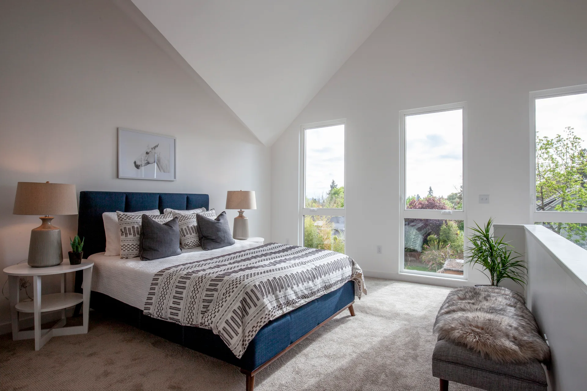 A contemporary bedroom with a blue upholstered bed, patterned textiles, round nightstands, a faux fur bench, and large windows under a vaulted ceiling.
