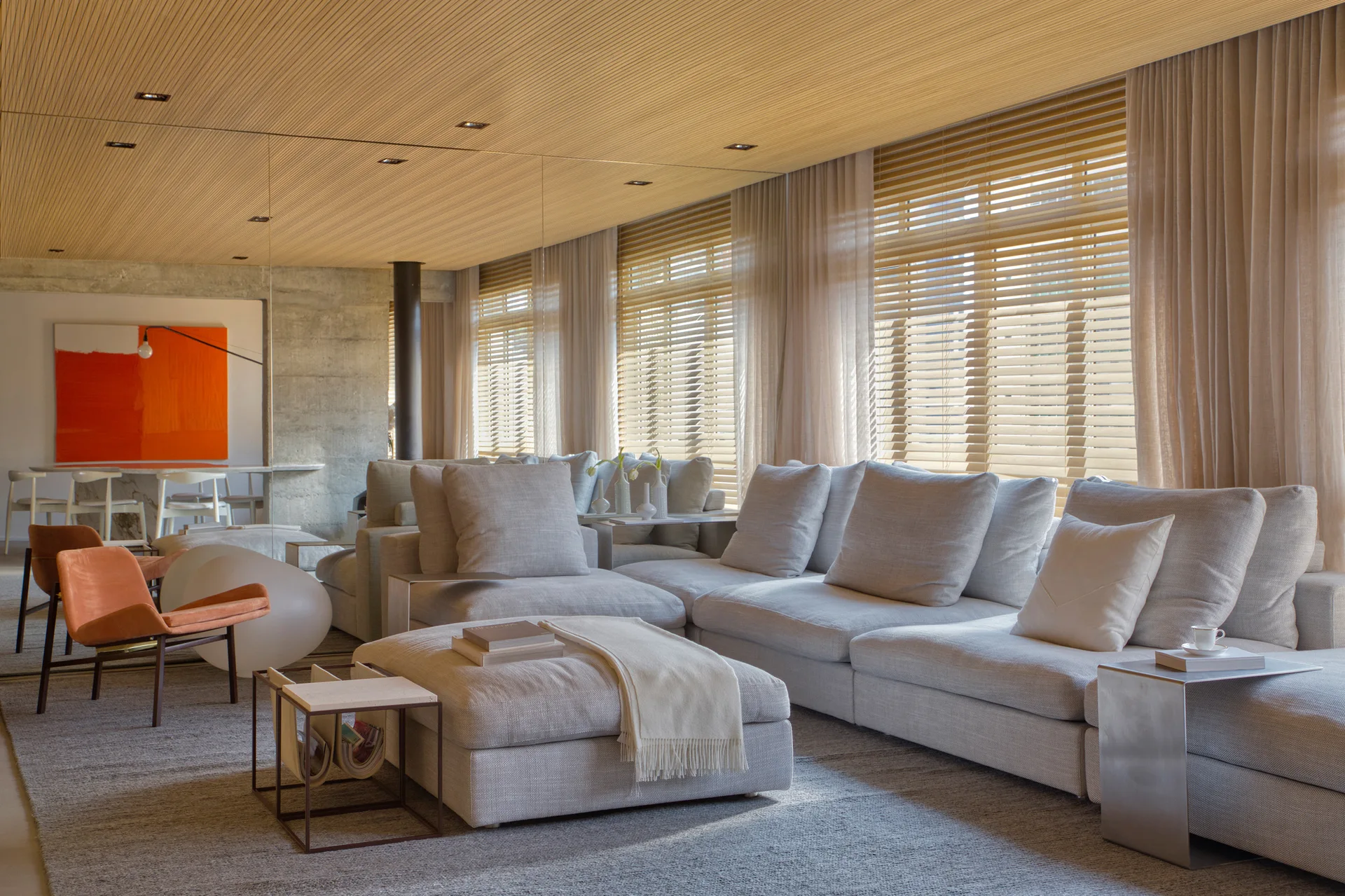 A modern living room featuring a large grey sectional sofa, matching ottoman, sheer drapes, and a linear light wood slat ceiling.
