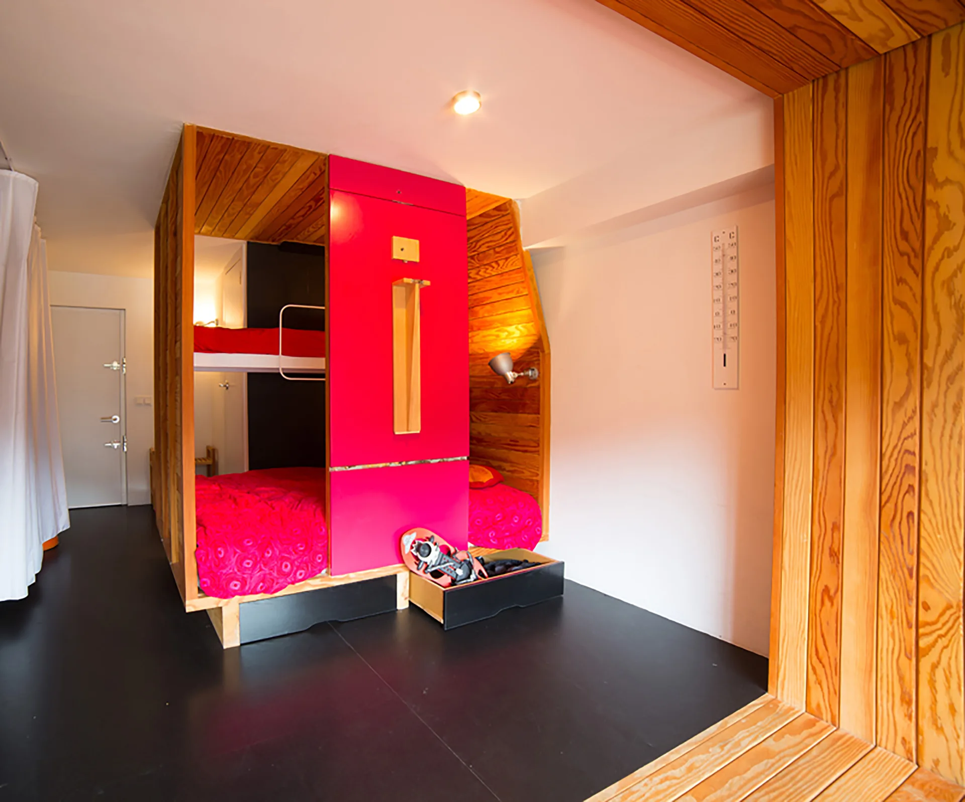 A modern bedroom features custom wood bunk beds with a bright pink central panel, matching pink bedding, and a black floor.