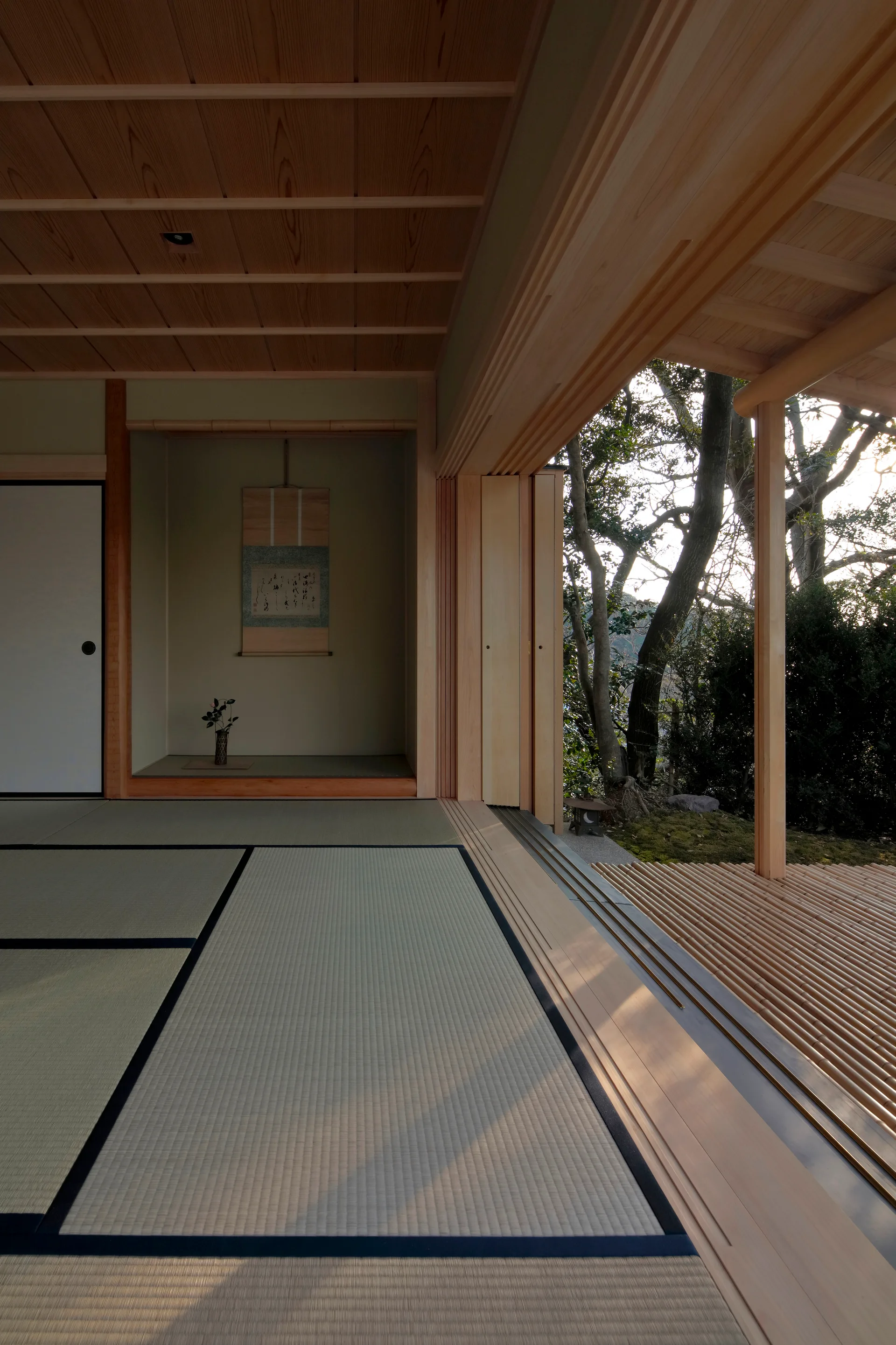 A traditional Japanese room features tatami flooring, a recessed tokonoma alcove with a scroll, and open sliding doors leading to a wooden slatted deck.