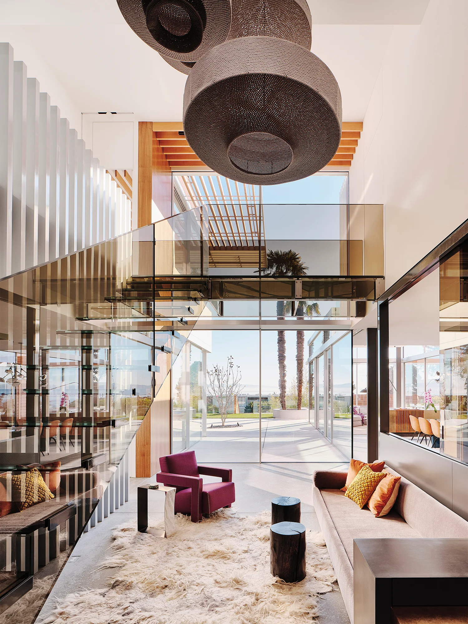 A high-ceilinged modern living room features a glass staircase, a purple armchair on a shaggy rug, and large windows looking out to a sunlit patio.