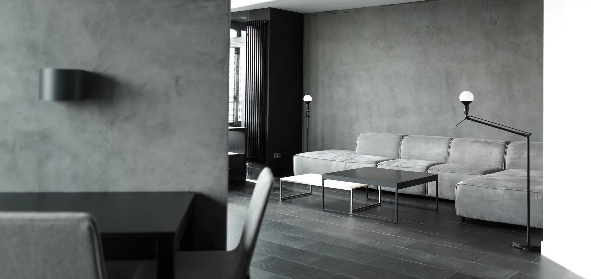 A minimalist grey living room features a low-profile sectional sofa, nested black and white coffee tables, and textured concrete-style walls.