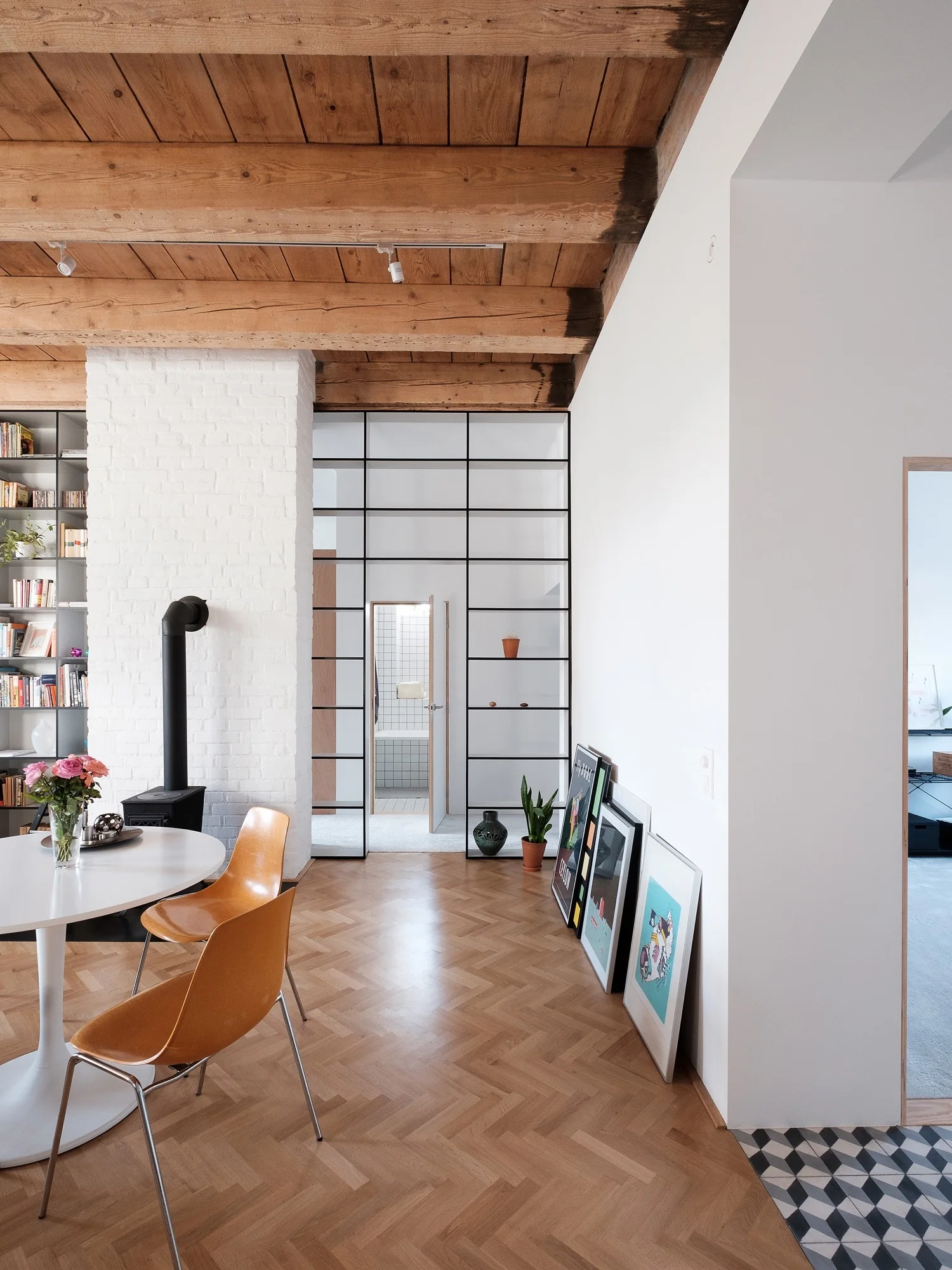 A dining room features herringbone wood floors, a round white pedestal table with orange chairs, a white brick fireplace, and exposed wood ceiling beams.