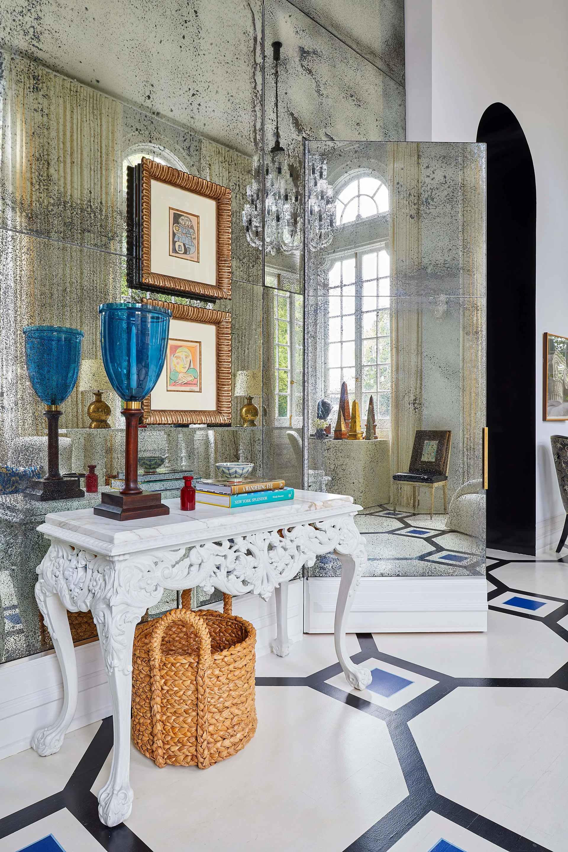 A carved white console table sits against an antique mirrored wall above a black and white geometric painted floor in an entryway.