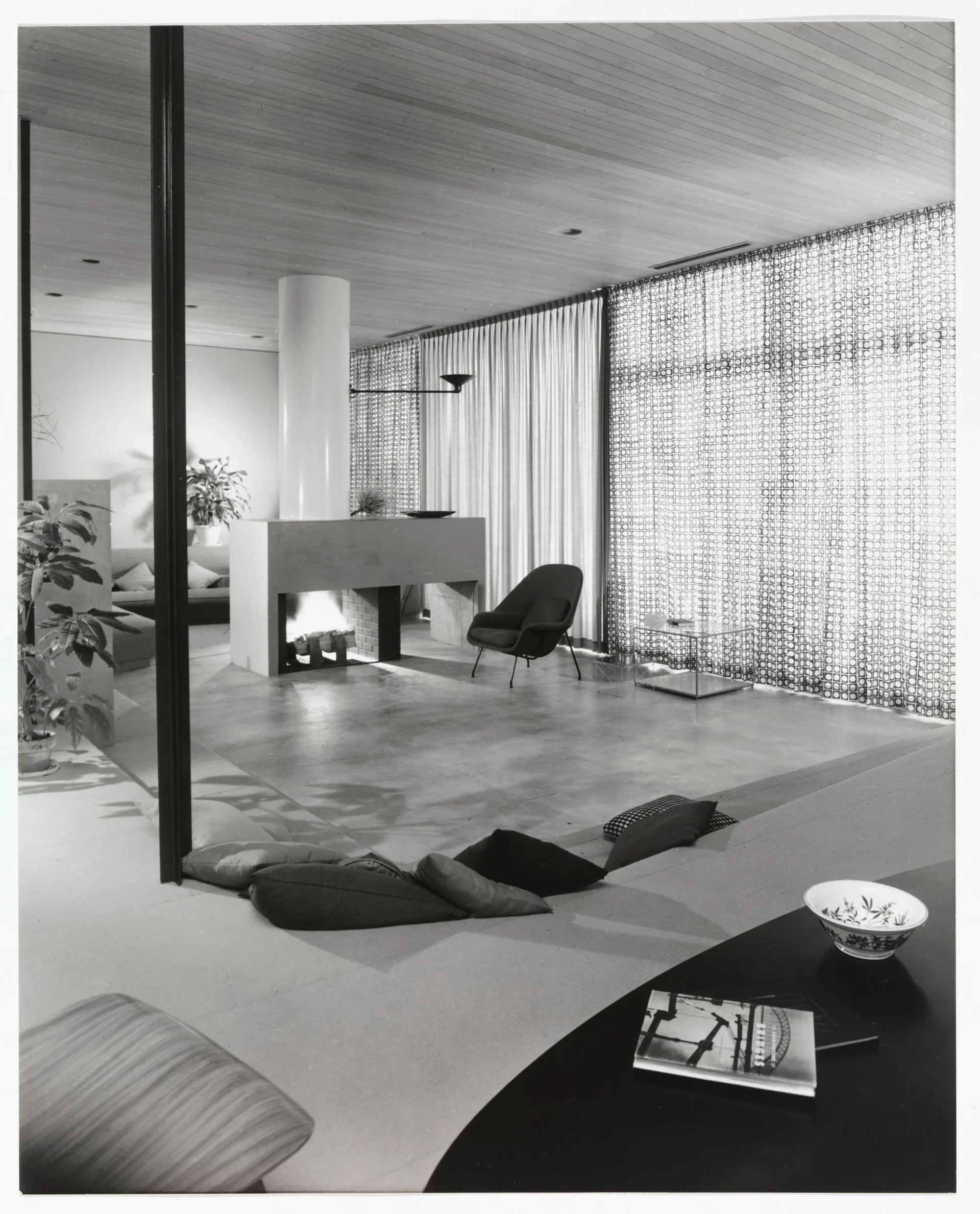 A mid-century modern living room features a sunken conversation pit, a central fireplace, a wood plank ceiling, and geometric patterned curtains.
