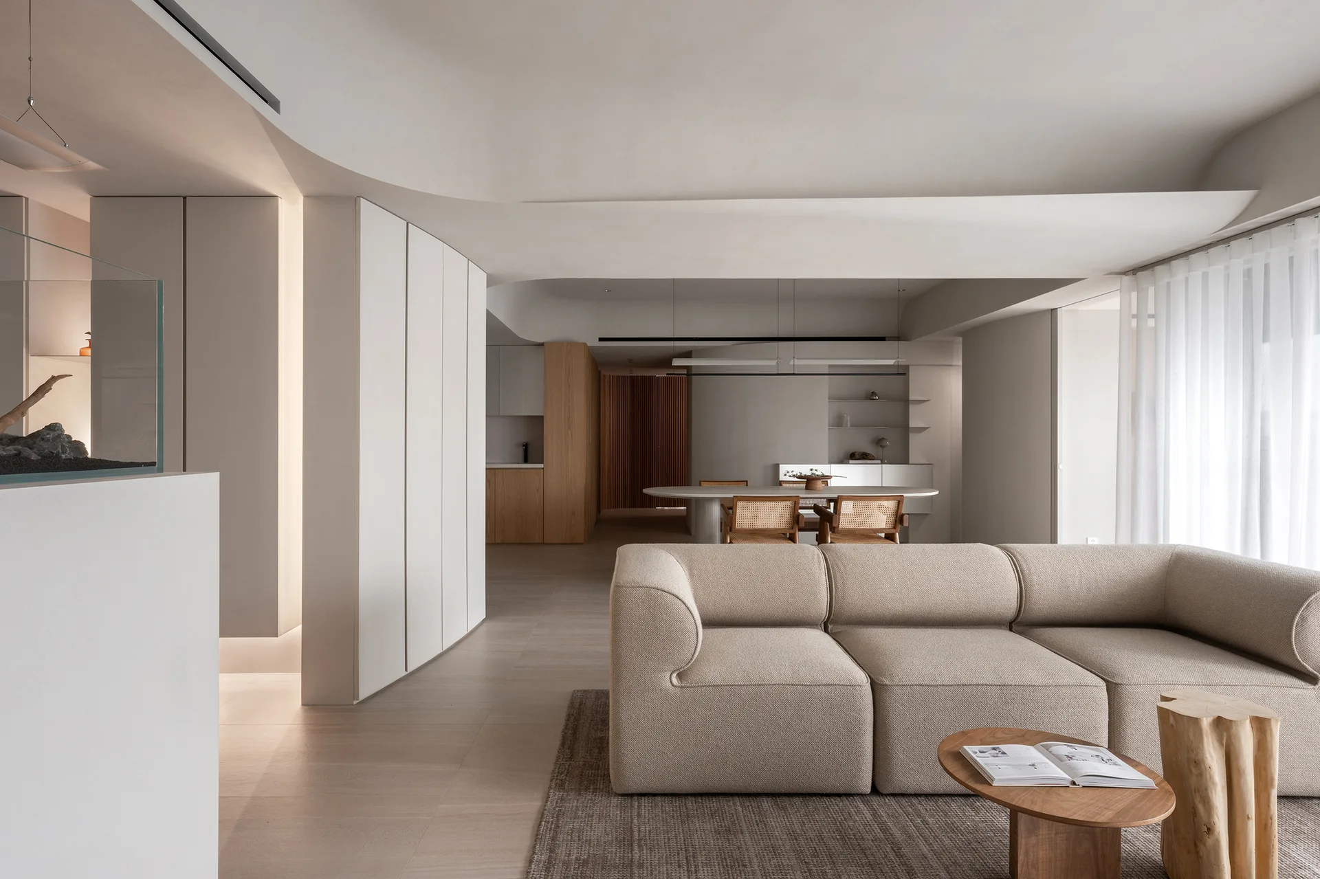 A minimalist living room with a beige modular sofa, wooden coffee table, and an open dining area beneath a curved ceiling soffit.