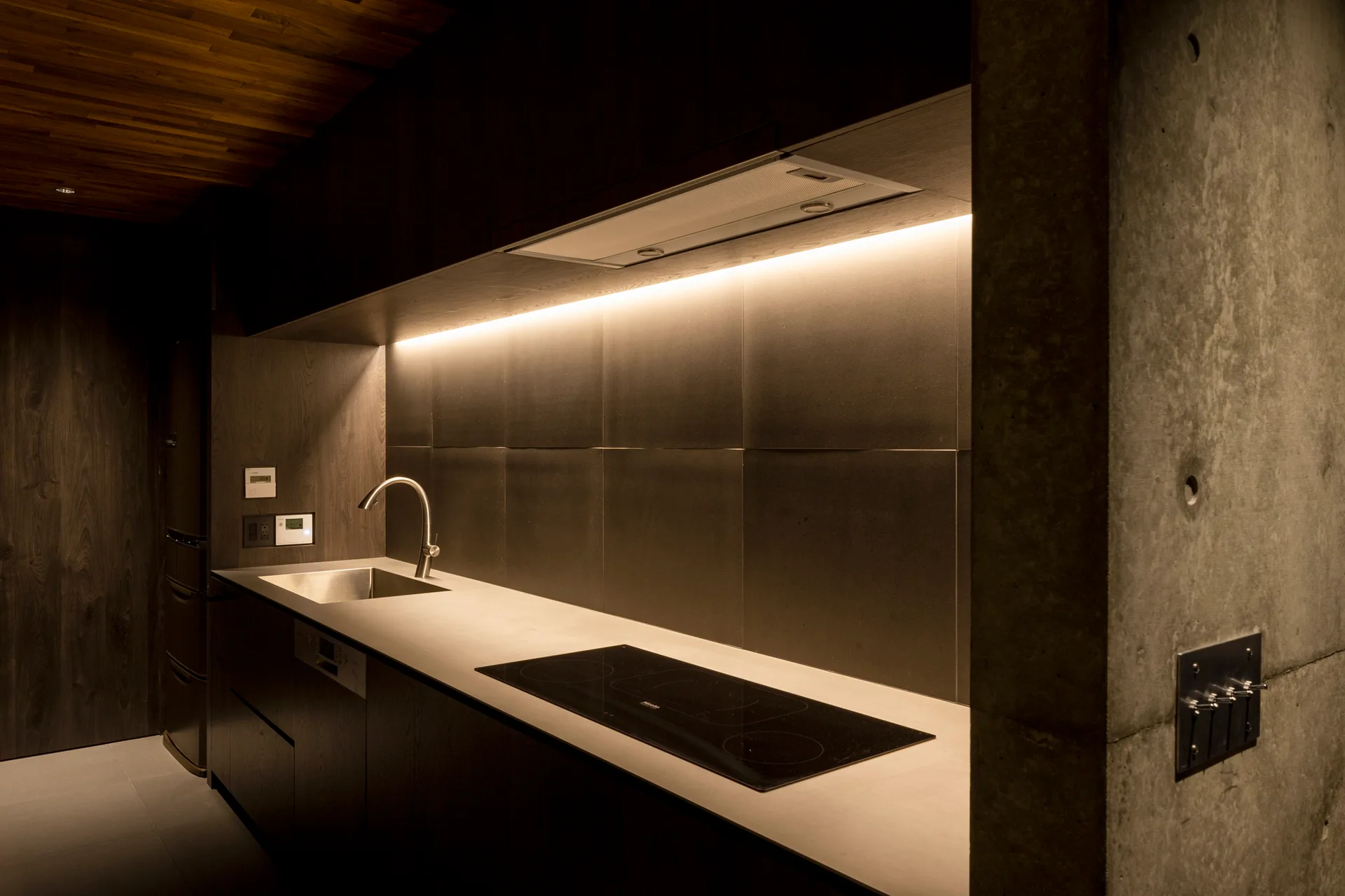 A dark modern kitchen features wood cabinets, a concrete wall pillar, an integrated cooktop, and warm under-cabinet strip lighting.