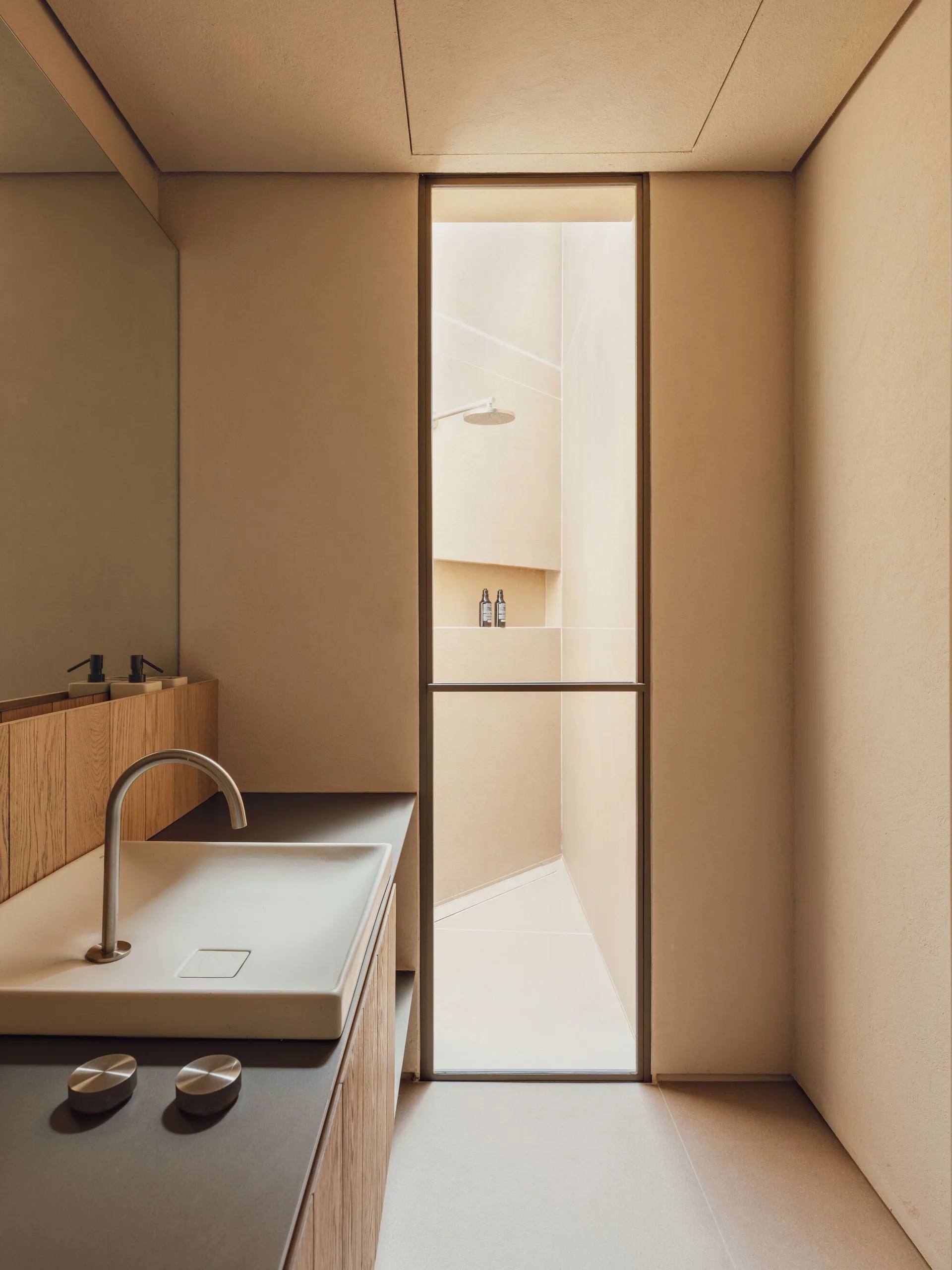 A minimalist bathroom with a light wood vanity, white rectangular sink basin, and a full-height glass door leading into a plaster shower.