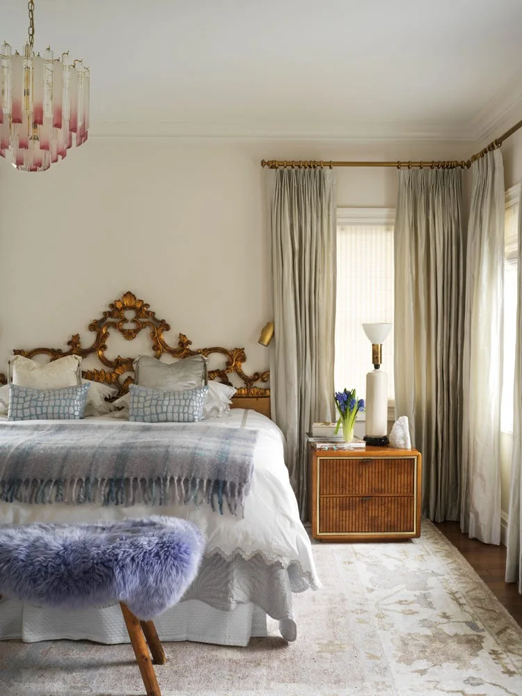 A bedroom featuring an ornate gold headboard on a bed with white bedding, alongside a fluted wood nightstand and a purple faux fur bench.