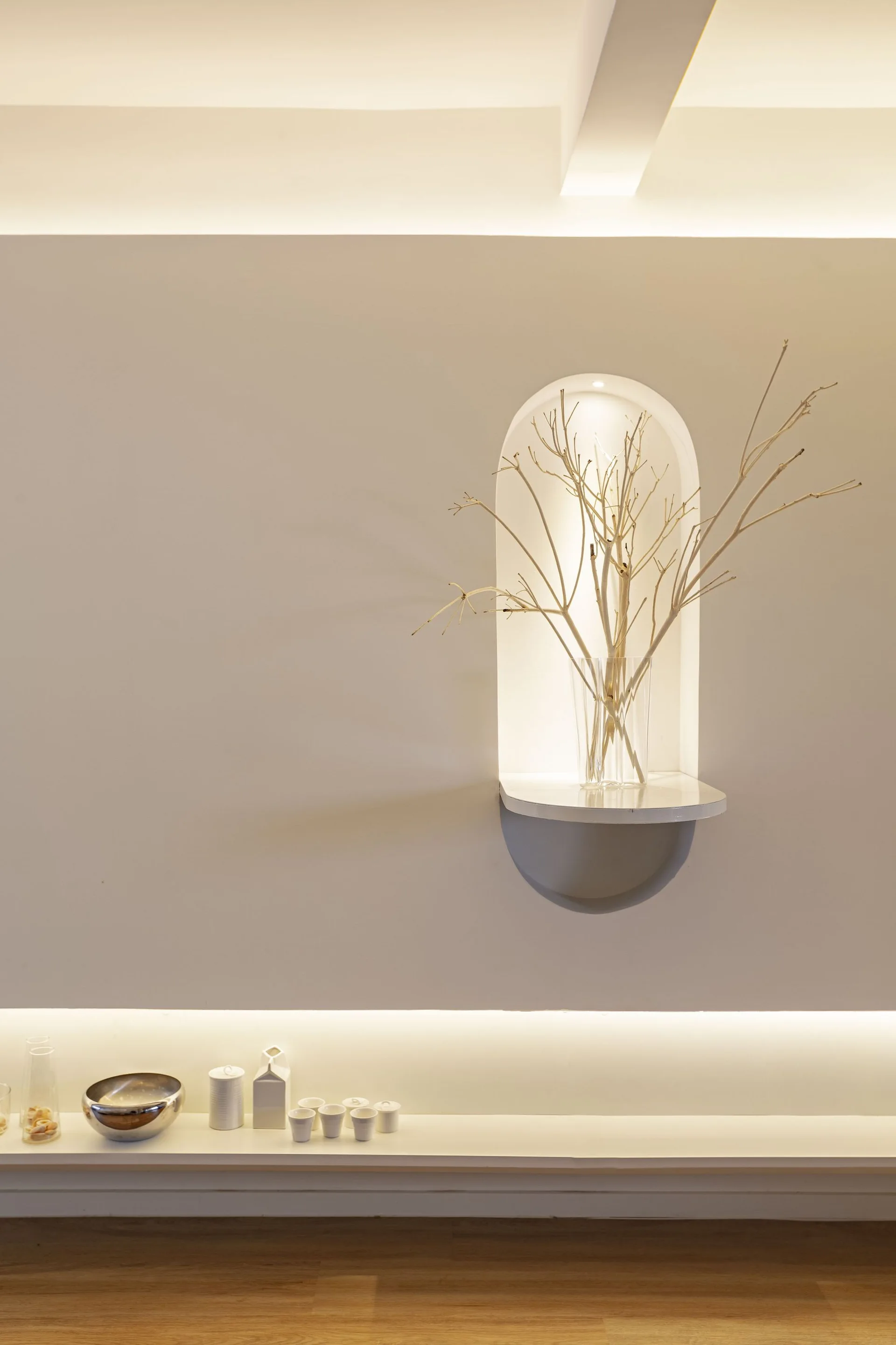 A minimalist off-white wall features a backlit arched niche with branches in a glass vase above a low display shelf with small ceramic accessories.