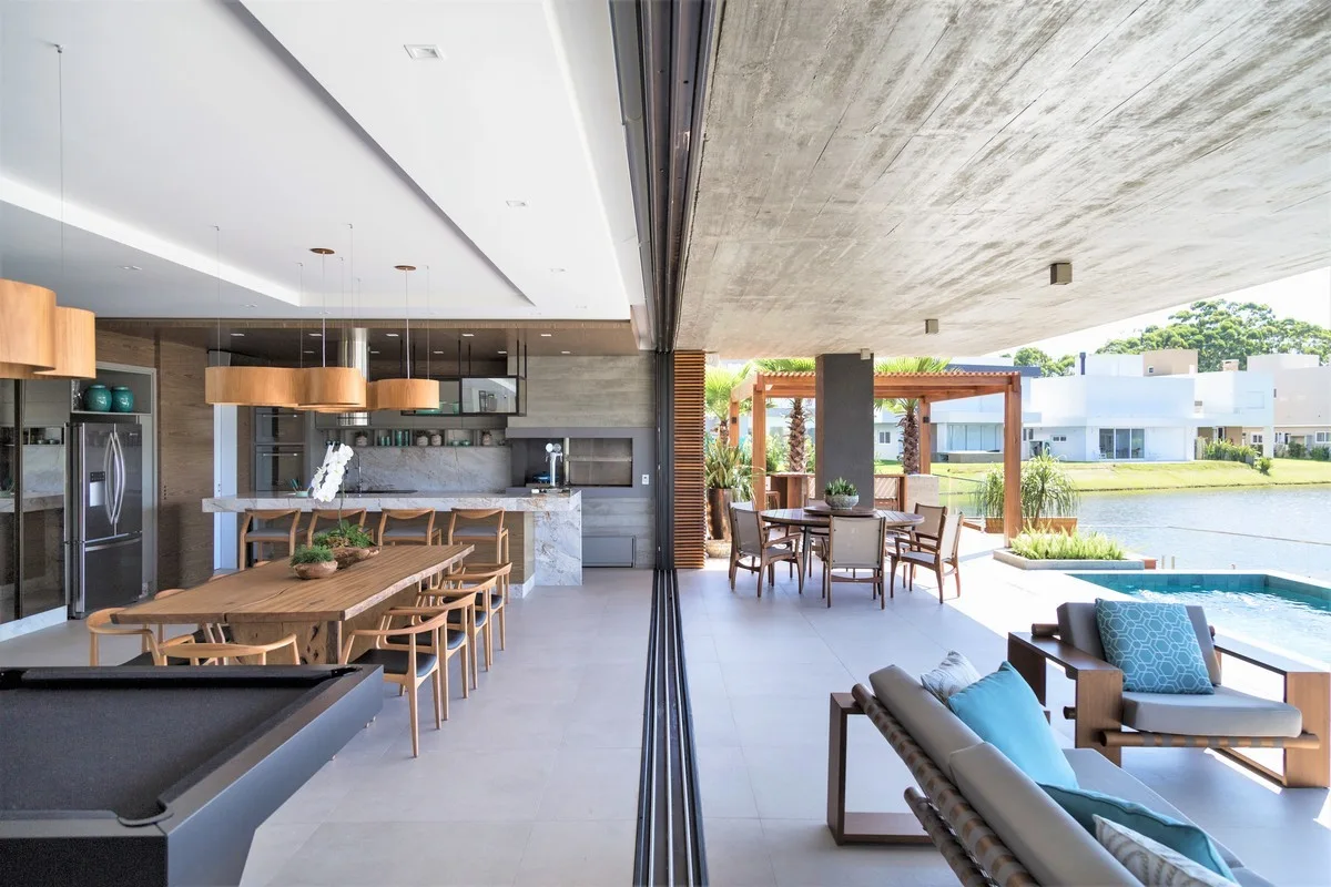 An open-concept kitchen and wooden dining table merge with a concrete-ceiling patio and outdoor pool via sliding glass doors.