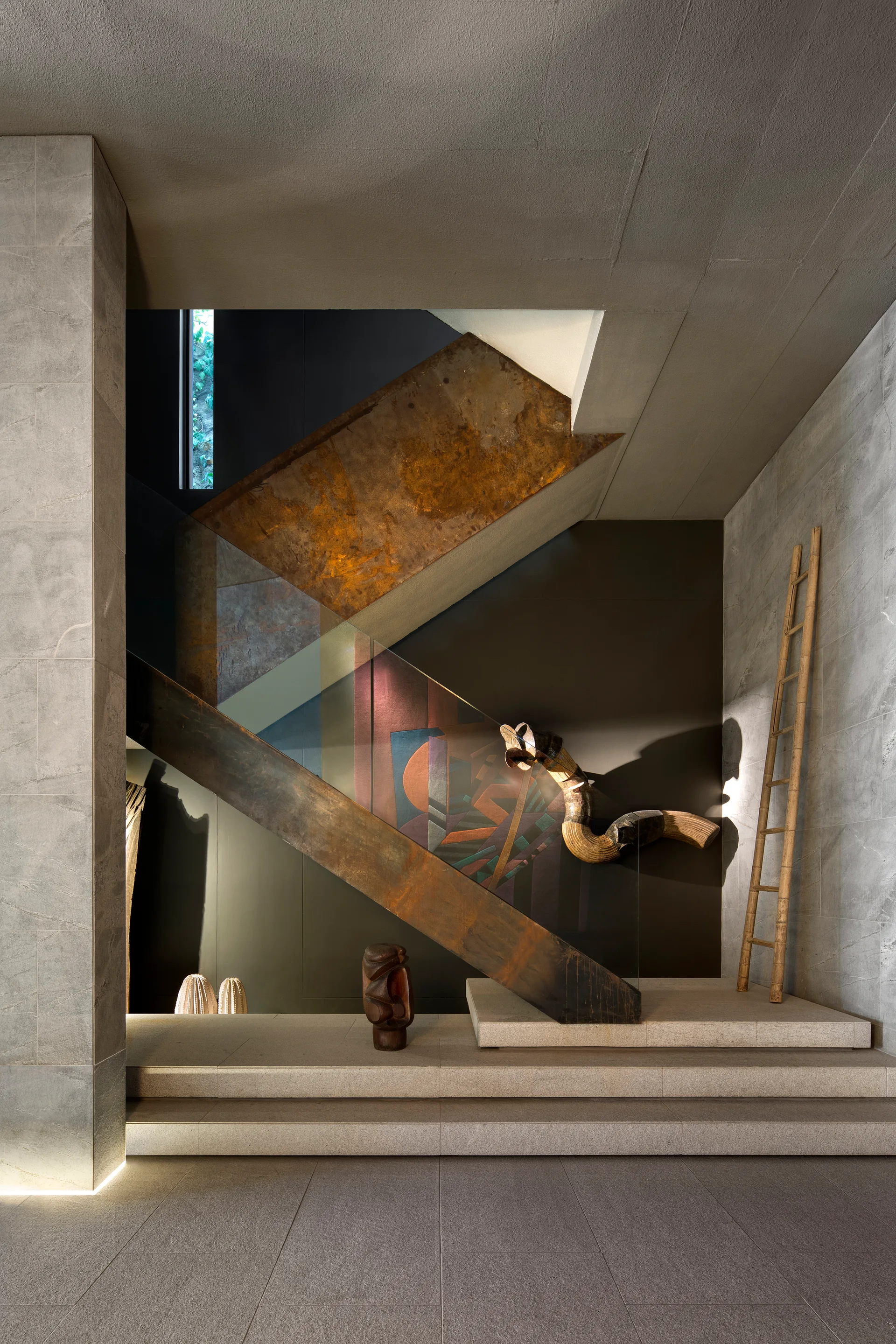 An industrial-style staircase featuring a rusted metal stringer and clear glass railing set against a dark wall with a geometric mural.
