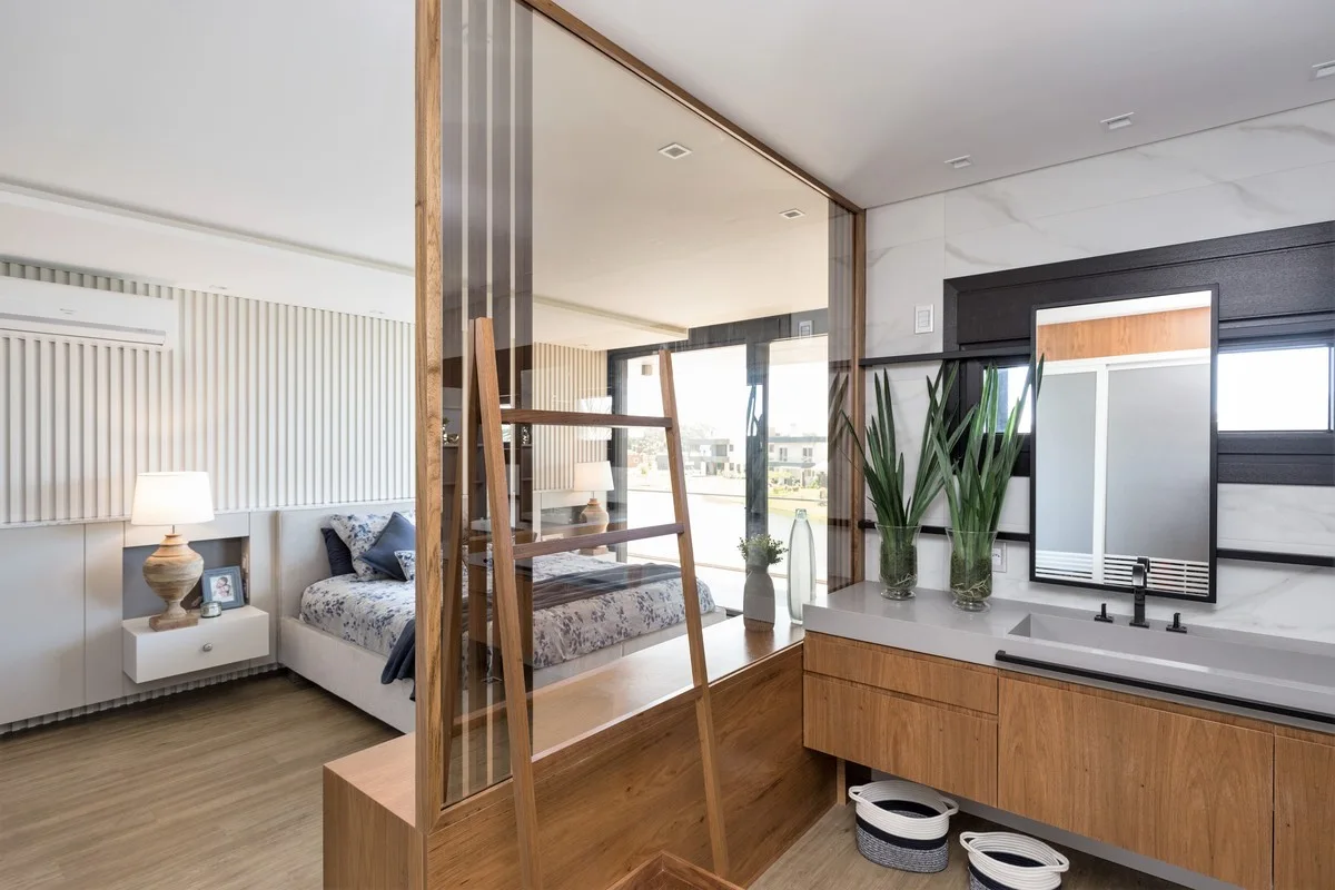 A contemporary bedroom and ensuite bathroom separated by a wood and glass partition, featuring a floating wood vanity and a white slatted wall.