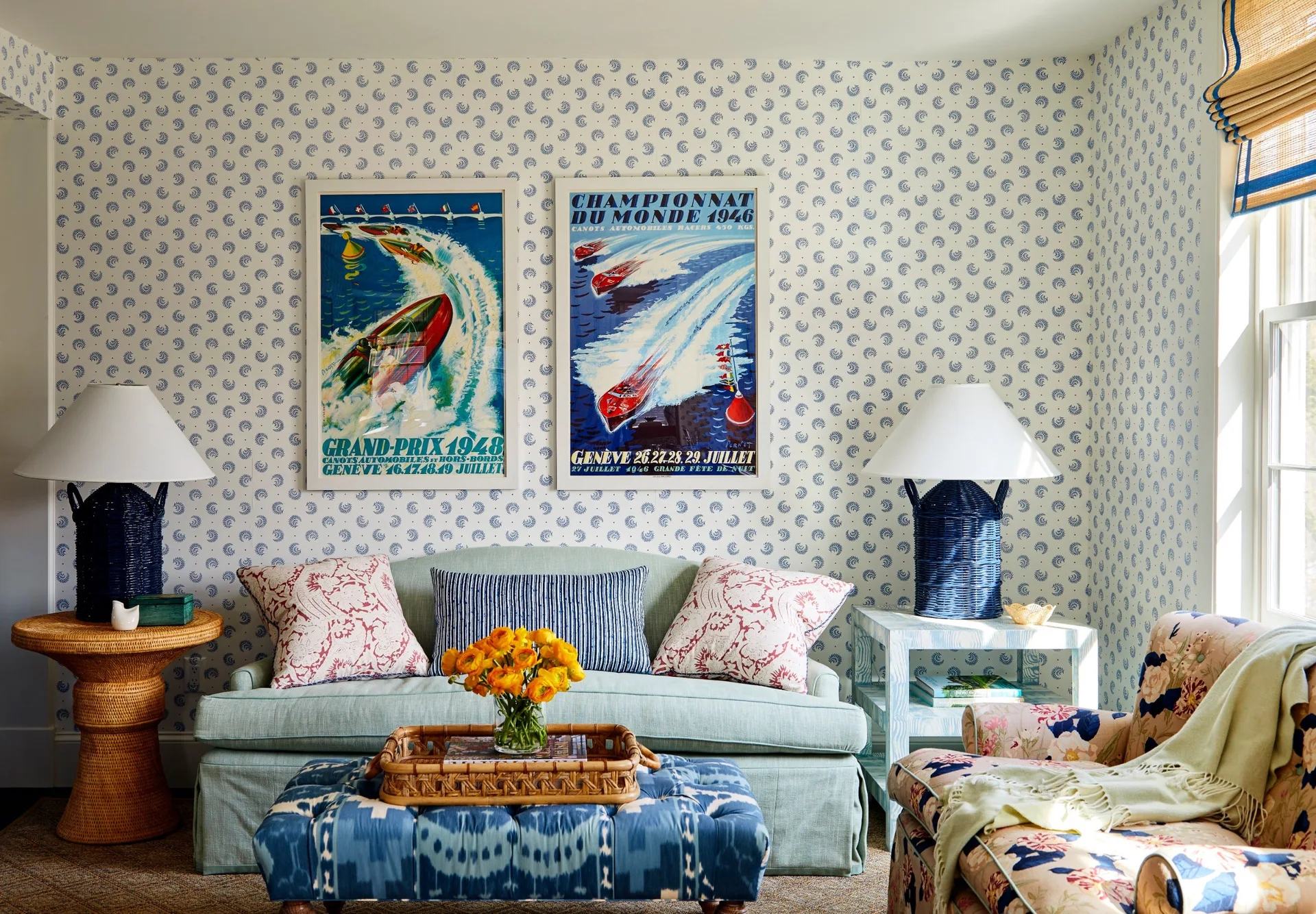 A light blue sofa and patterned ottoman sit against small-print wallpaper under two framed vintage boating posters flanked by blue wicker table lamps.