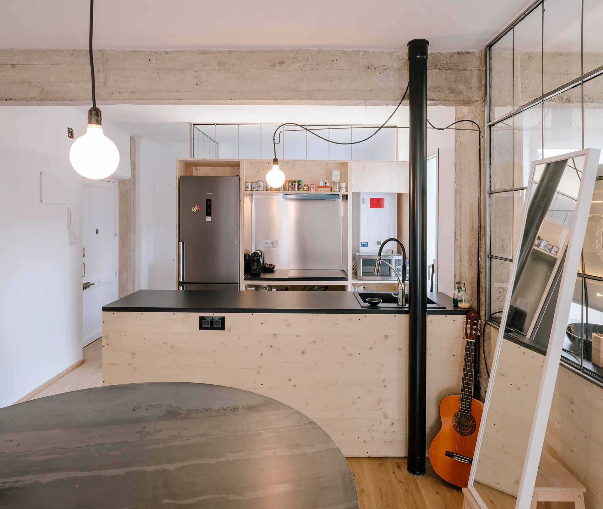 A modern kitchen featuring a plywood island with a black countertop, exposed concrete ceiling beam, and industrial steel-framed window wall.