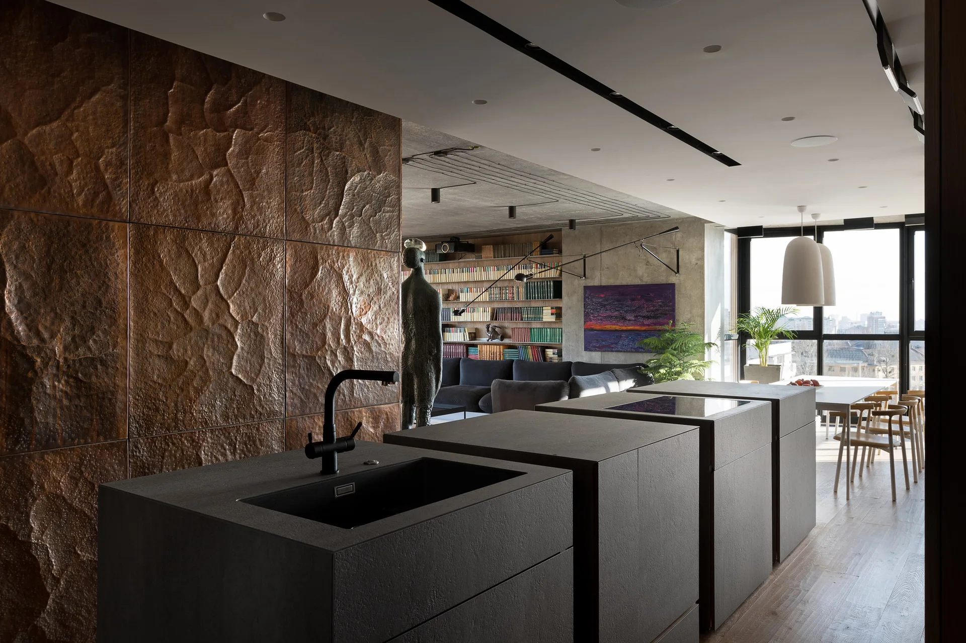 A modern open kitchen features dark gray island units and a textured copper wall, with a living room and dining area visible in the background.