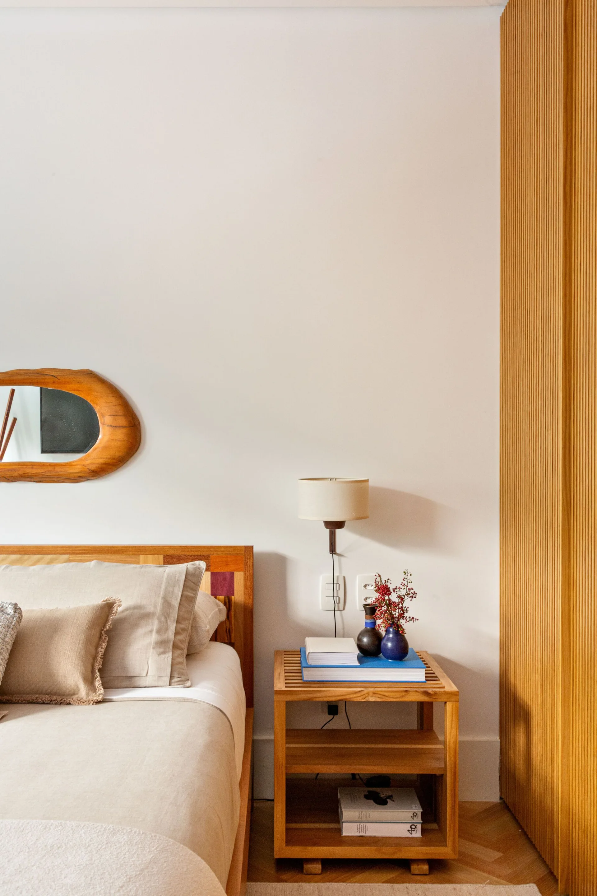 A bedroom corner features a wooden bed, a slatted wood nightstand with books and a vase, a wall-mounted lamp, and vertical wood paneling.