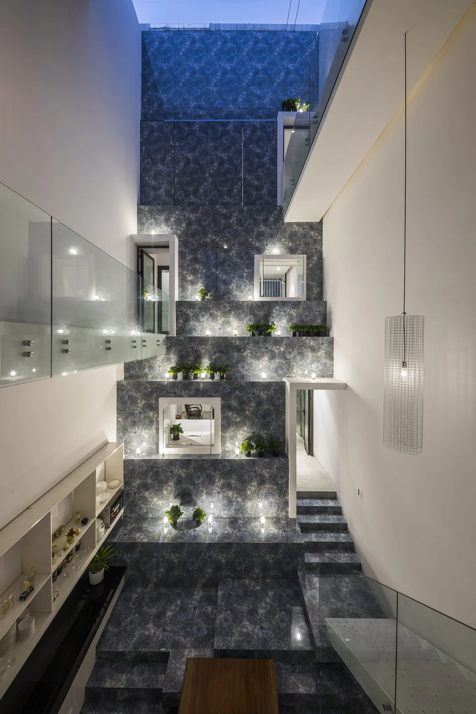 A modern multi-level stairwell featuring staggered blue patterned stone platforms, glass railings, and tiny integrated spotlights next to potted plants.