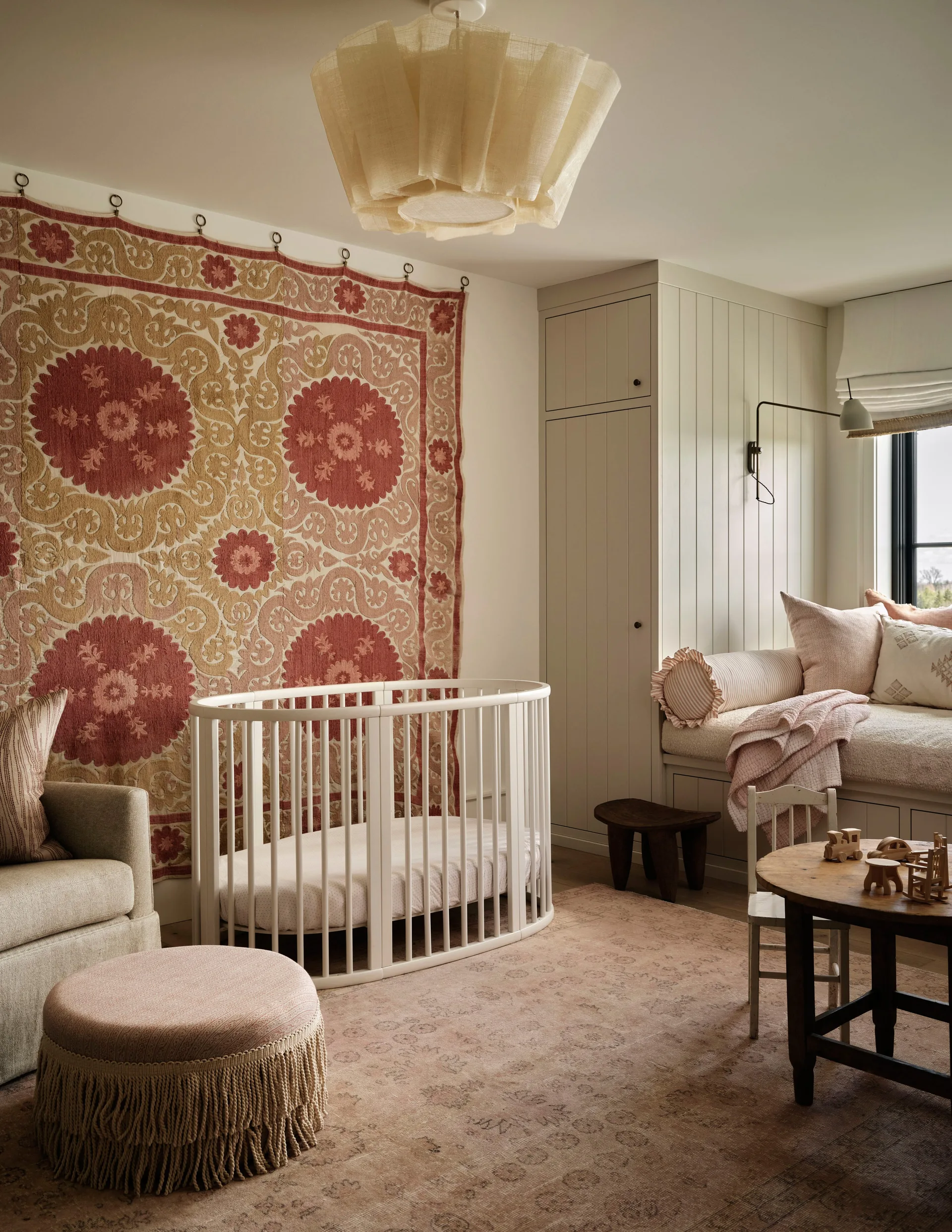 A nursery featuring a white oval wooden crib, a large terracotta and cream wall tapestry, and a built-in window bench with storage.