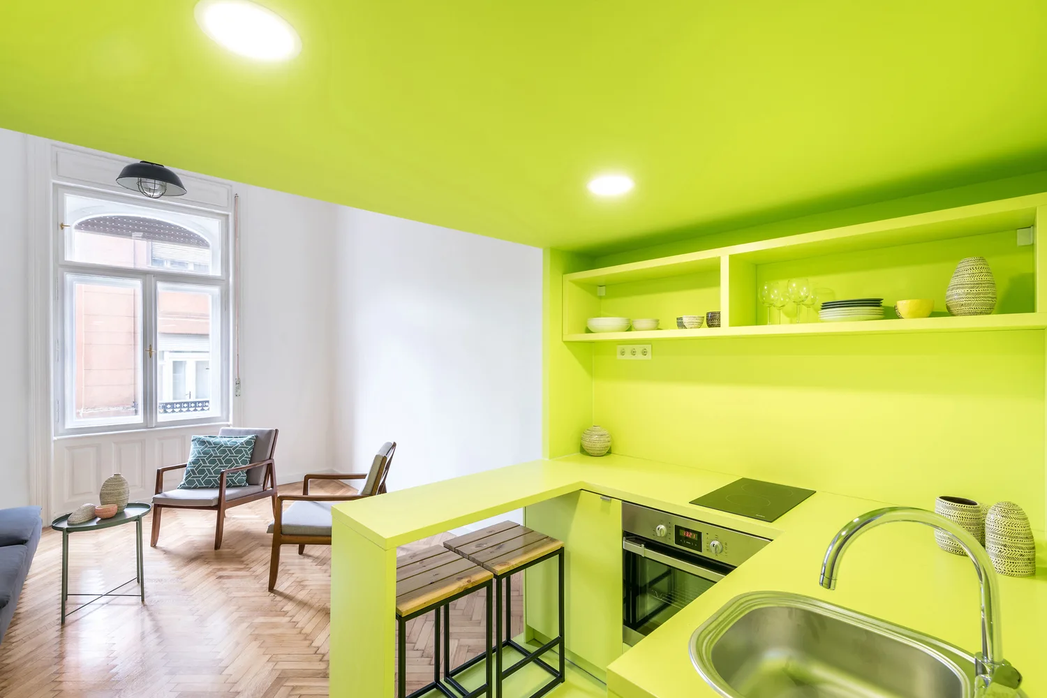 A compact kitchen block painted bright lime green sits adjacent to a white-walled living area with wood herringbone floors and grey armchairs.