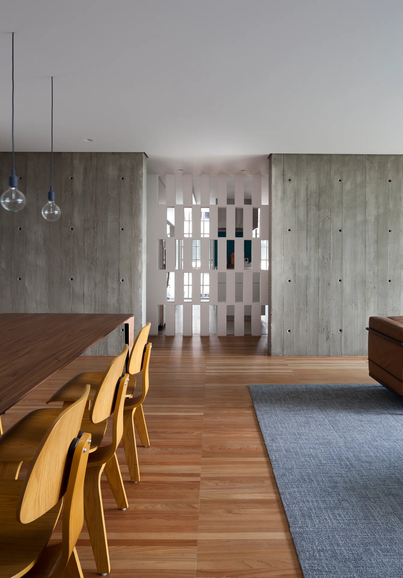 A modern dining room features wood furniture, exposed concrete walls, a grey rug, and a white geometric room divider in the background.