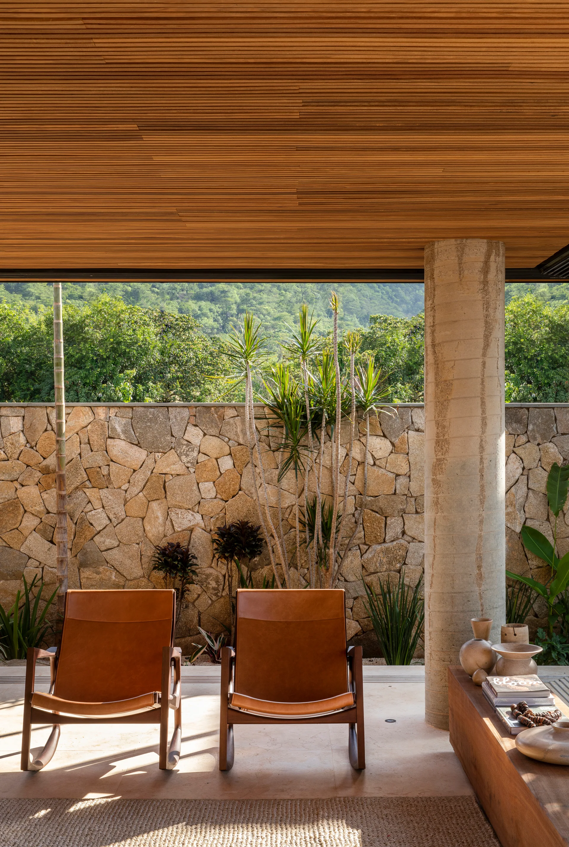 Two tan leather rocking chairs sit on a covered patio beneath a slatted wood ceiling, with a stacked stone wall and lush greenery in the background.