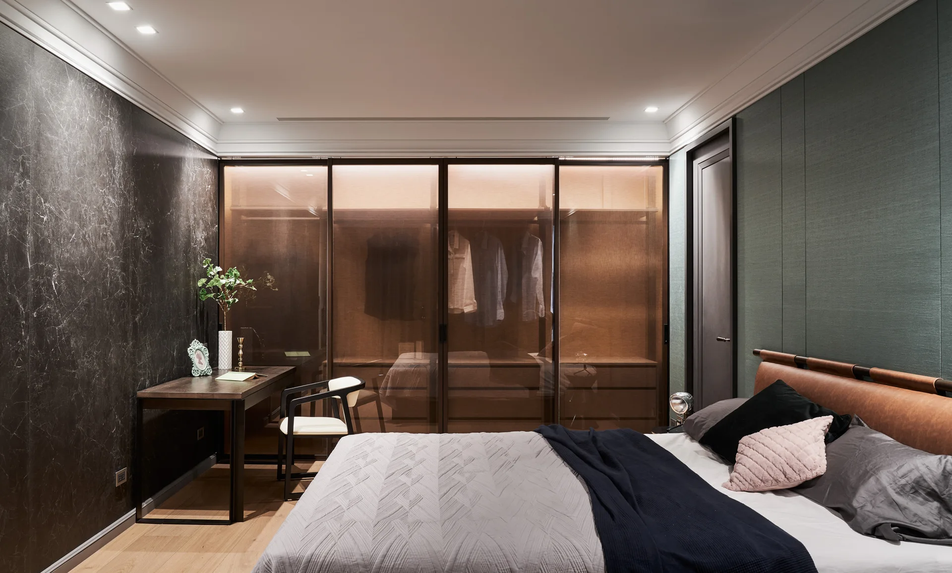 A modern bedroom features a bed with an upholstered headboard, a dark writing desk, and a built-in wardrobe with backlit tinted glass sliding doors.