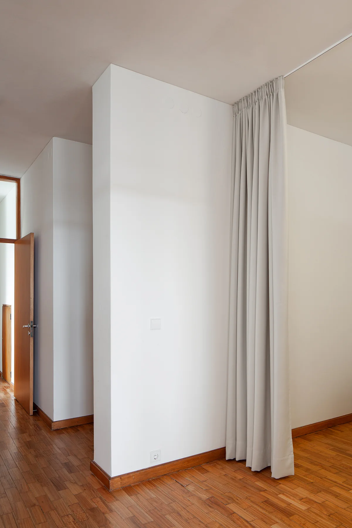 A beige floor-to-ceiling curtain hangs next to a white partition wall in a minimalist room with wood parquet flooring.