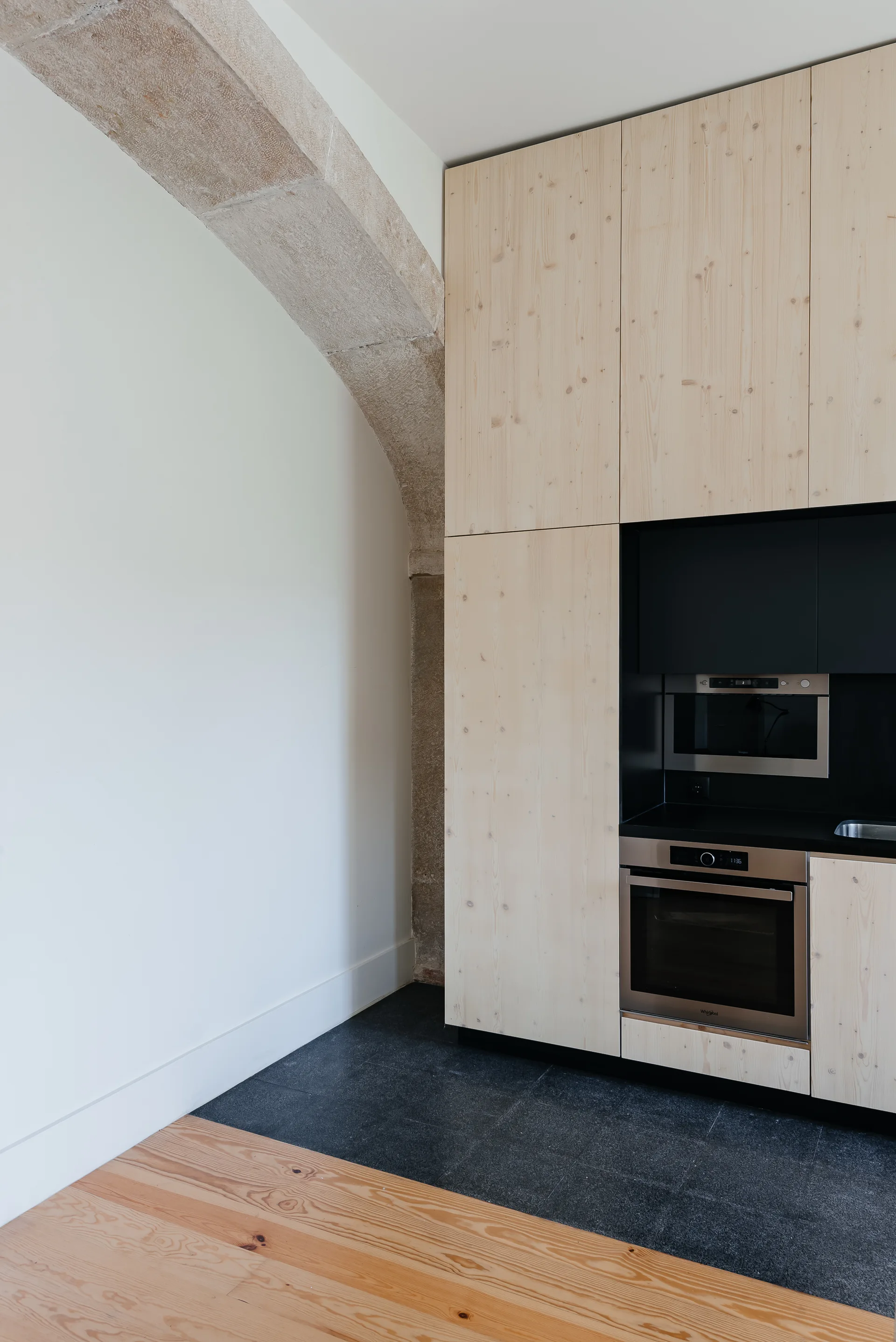 Flat-front light wood kitchen cabinets with integrated black appliances stand next to a historic textured stone archway on dark stone tile flooring.