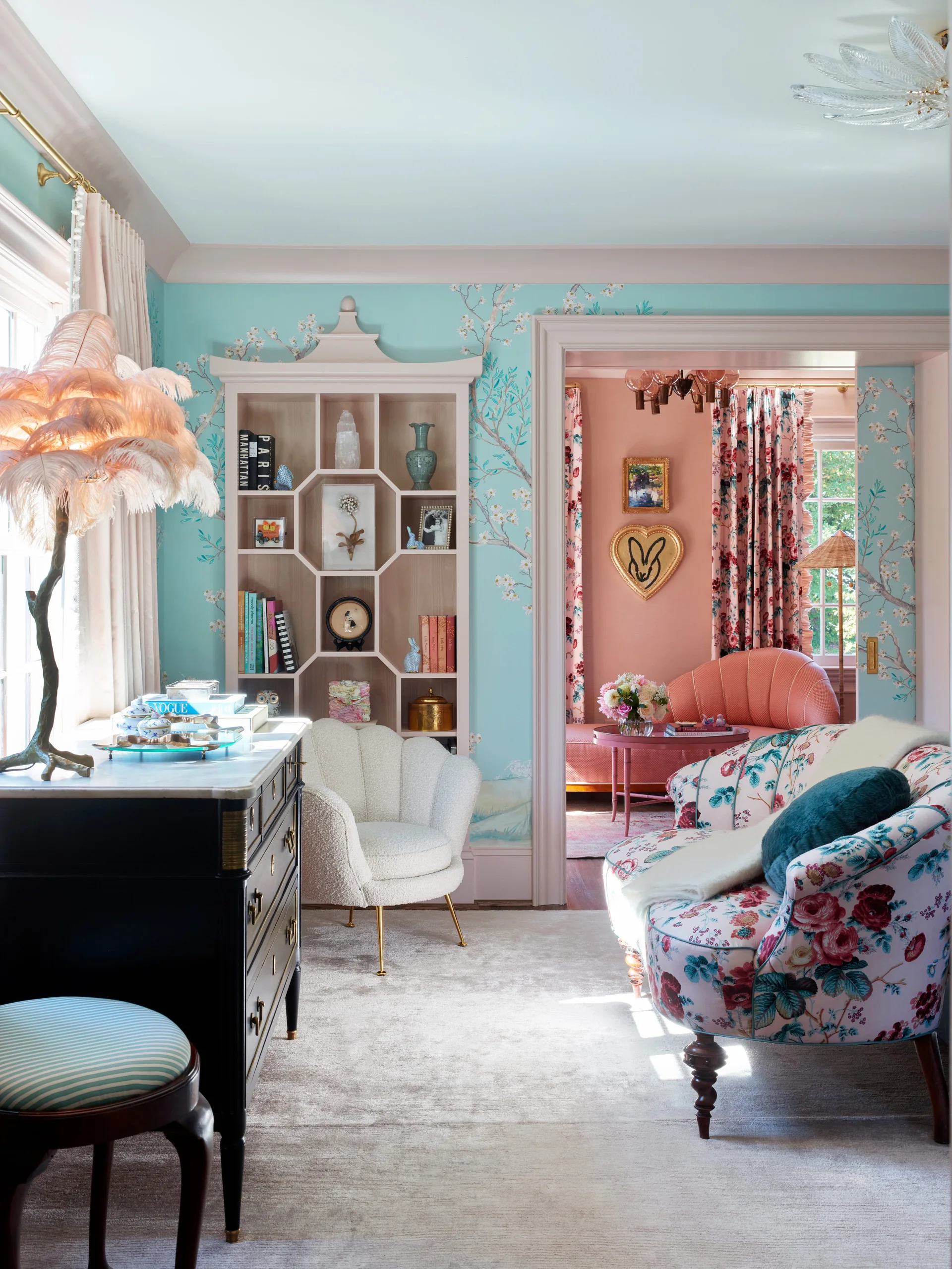 An eclectic living room with teal floral wallpaper, a light pink built-in bookcase, a white scalloped armchair, and a doorway opening to a pink room.