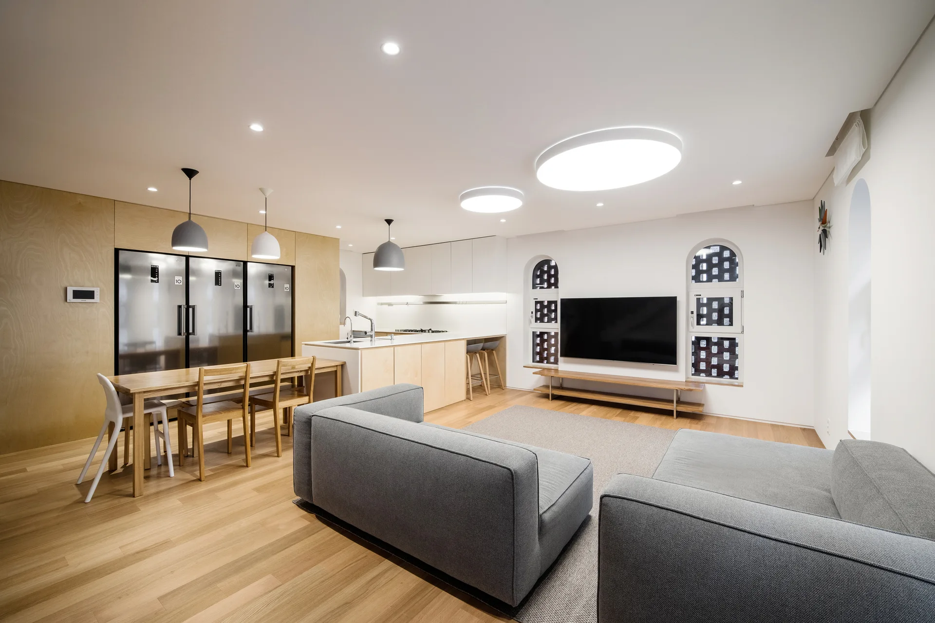 An open-plan living room and kitchen featuring a grey sectional sofa, light wood paneling, integrated appliances, and two small arched windows.