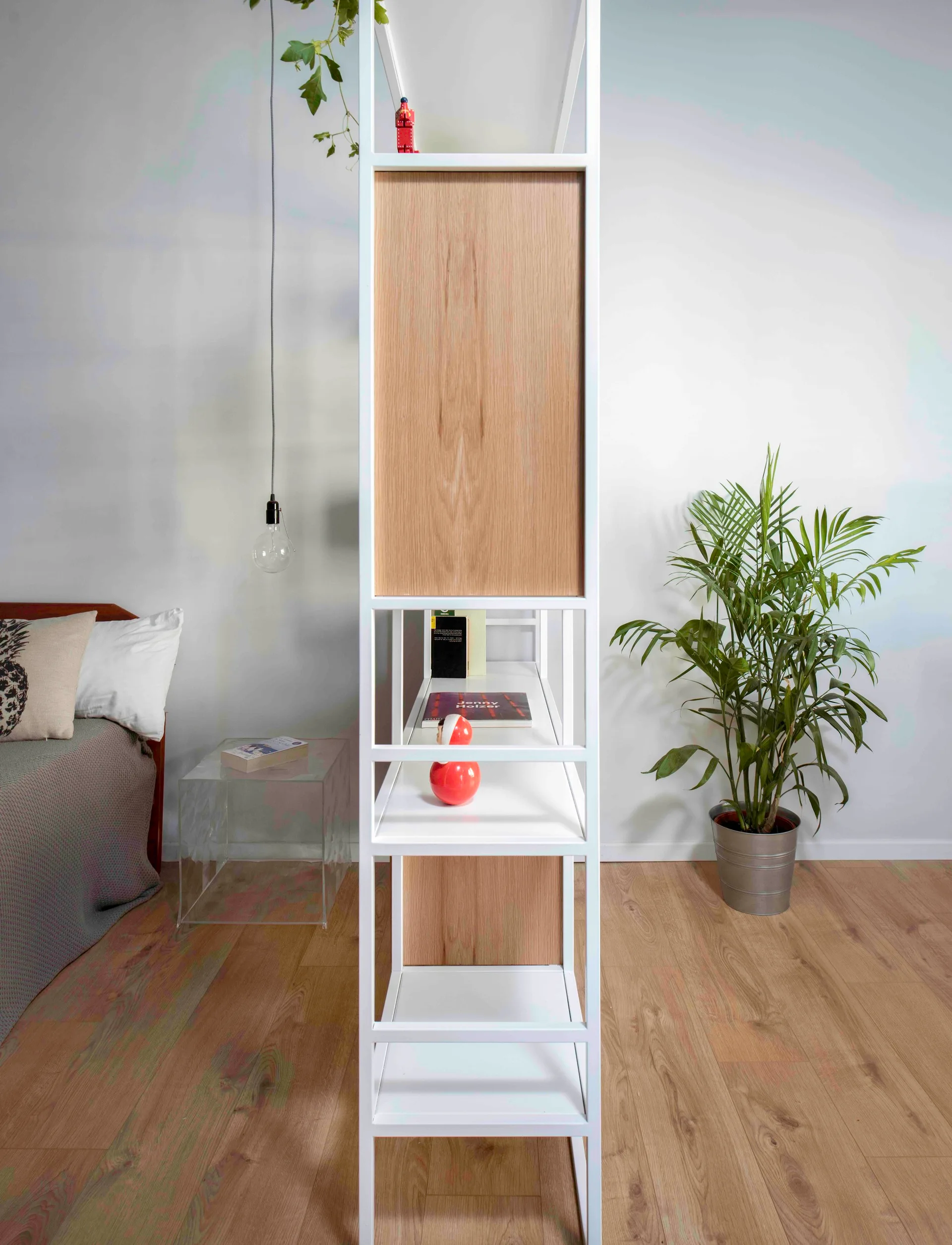 A tall white open-shelving room divider with light wood panels stands on wood floors in a minimalist bedroom near a clear acrylic side table and bed.